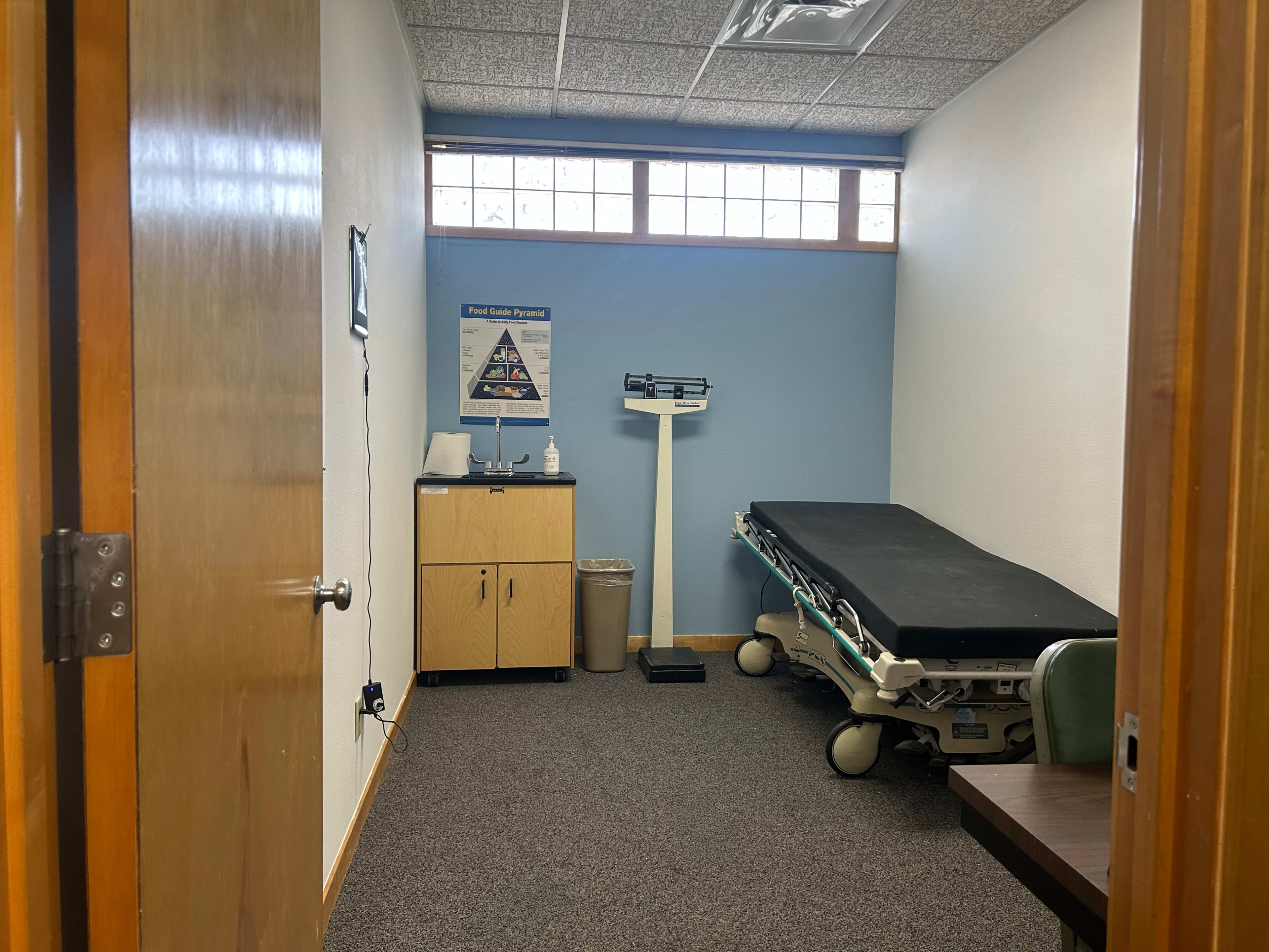 The image shows a small examination room with a medical examination table, a scale, a waste bin, and a cabinet against the wall.