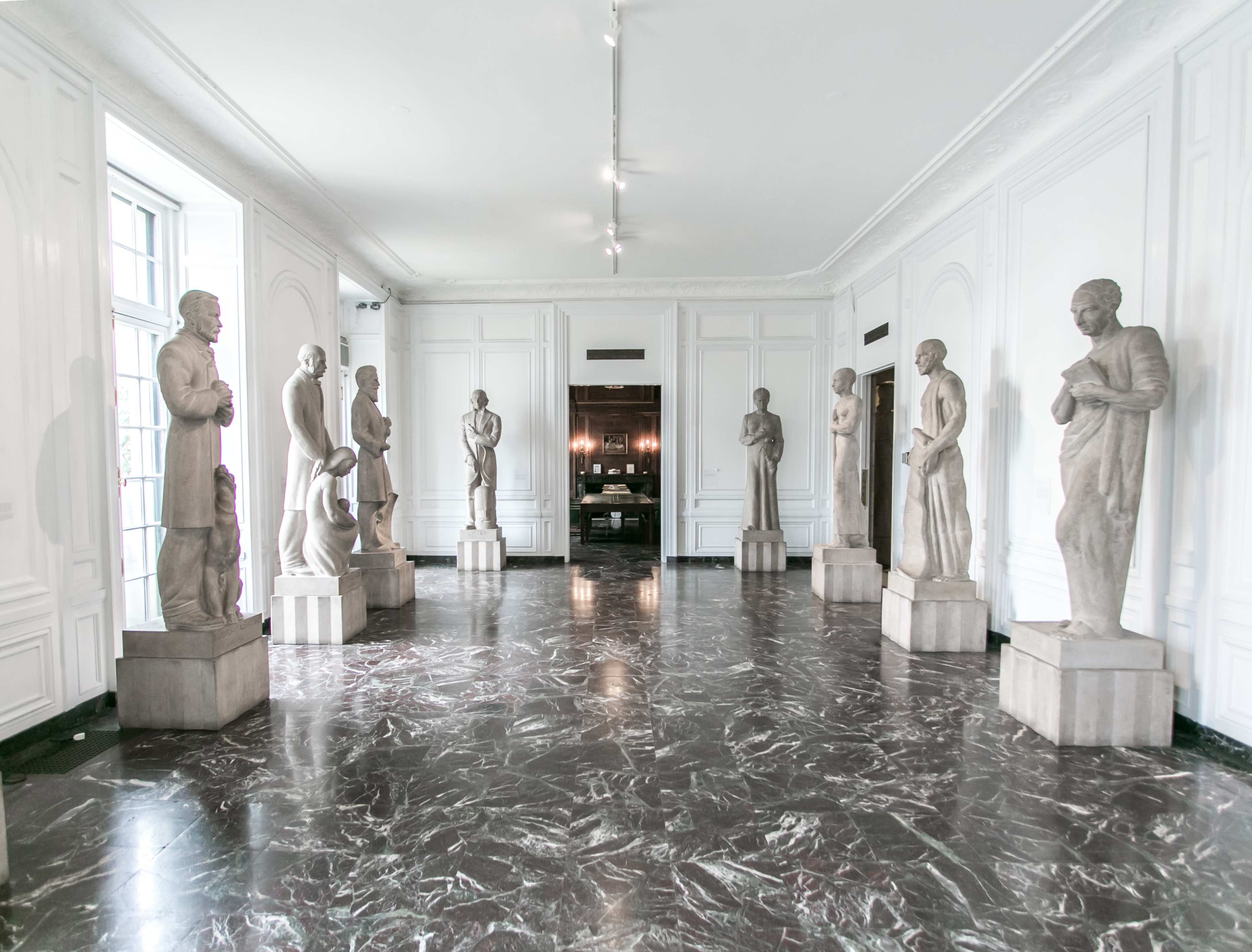 A marble-floored gallery with eight statues evenly spaced along the walls, illuminated by natural light from large windows.