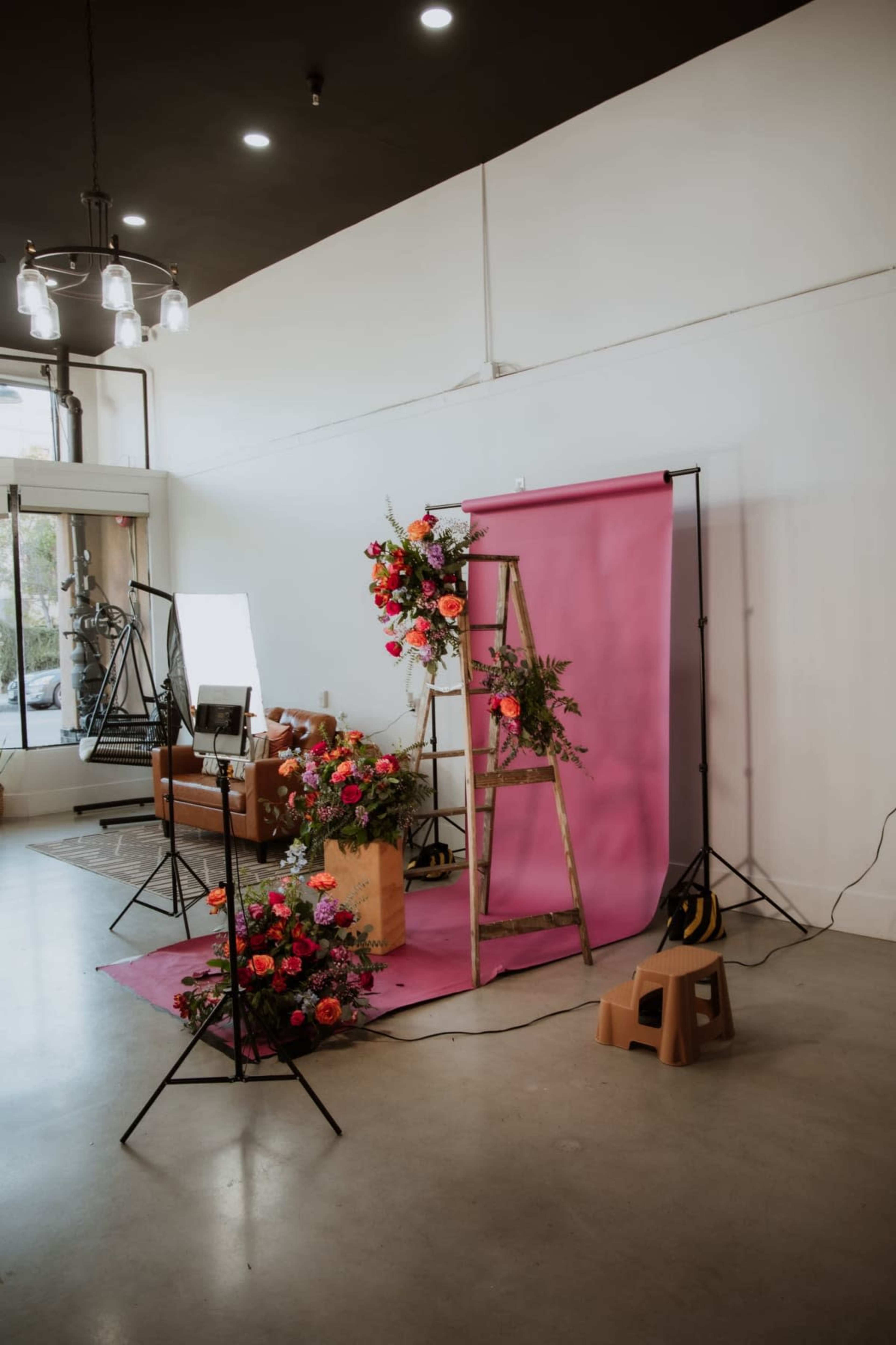 The image shows a photography setup featuring a pink backdrop, a wooden ladder adorned with flowers, and assorted lighting equipment in a studio space.