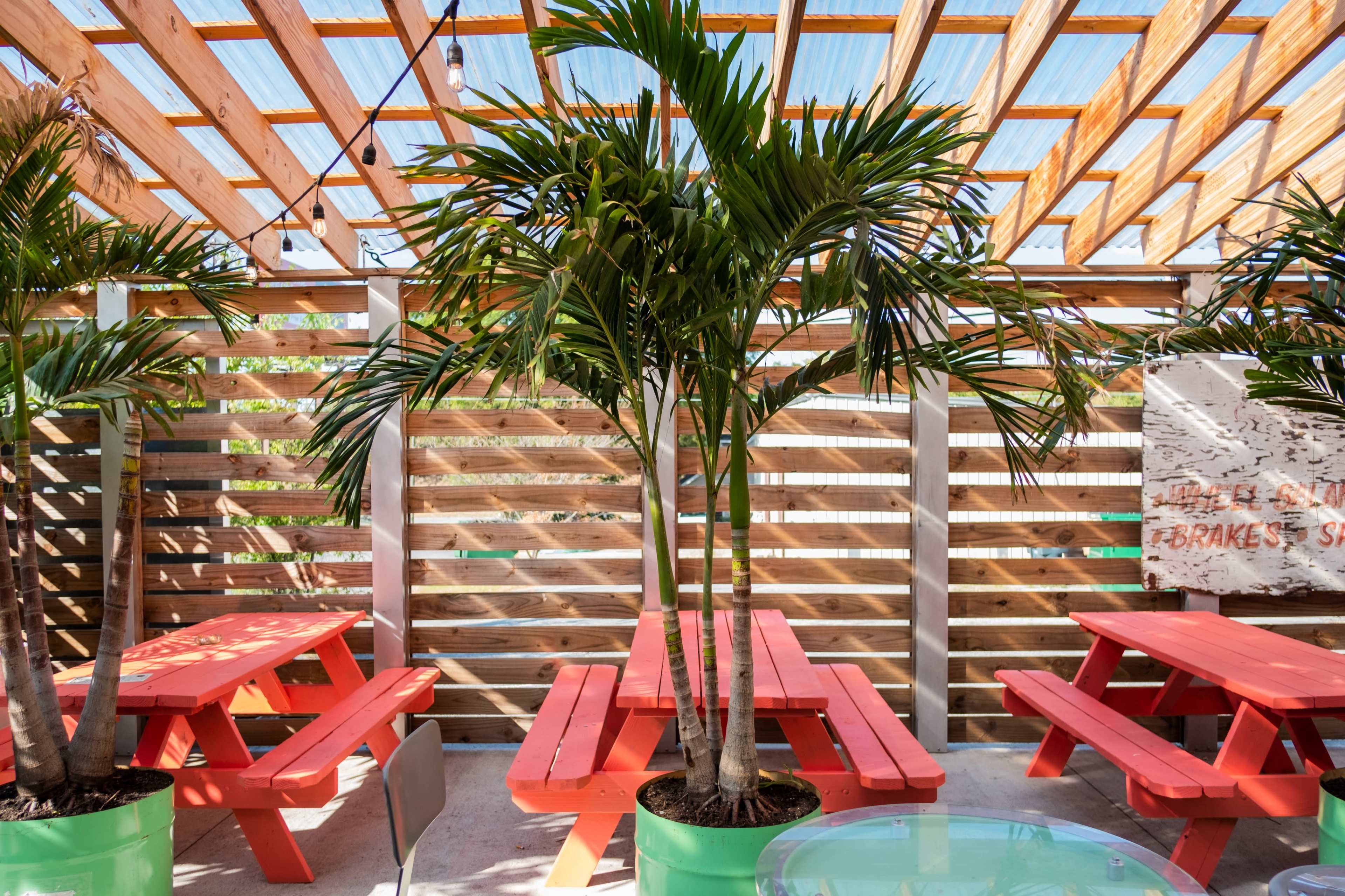 The image shows a bright outdoor dining area with wooden slatted walls, red picnic tables, and potted palm trees.