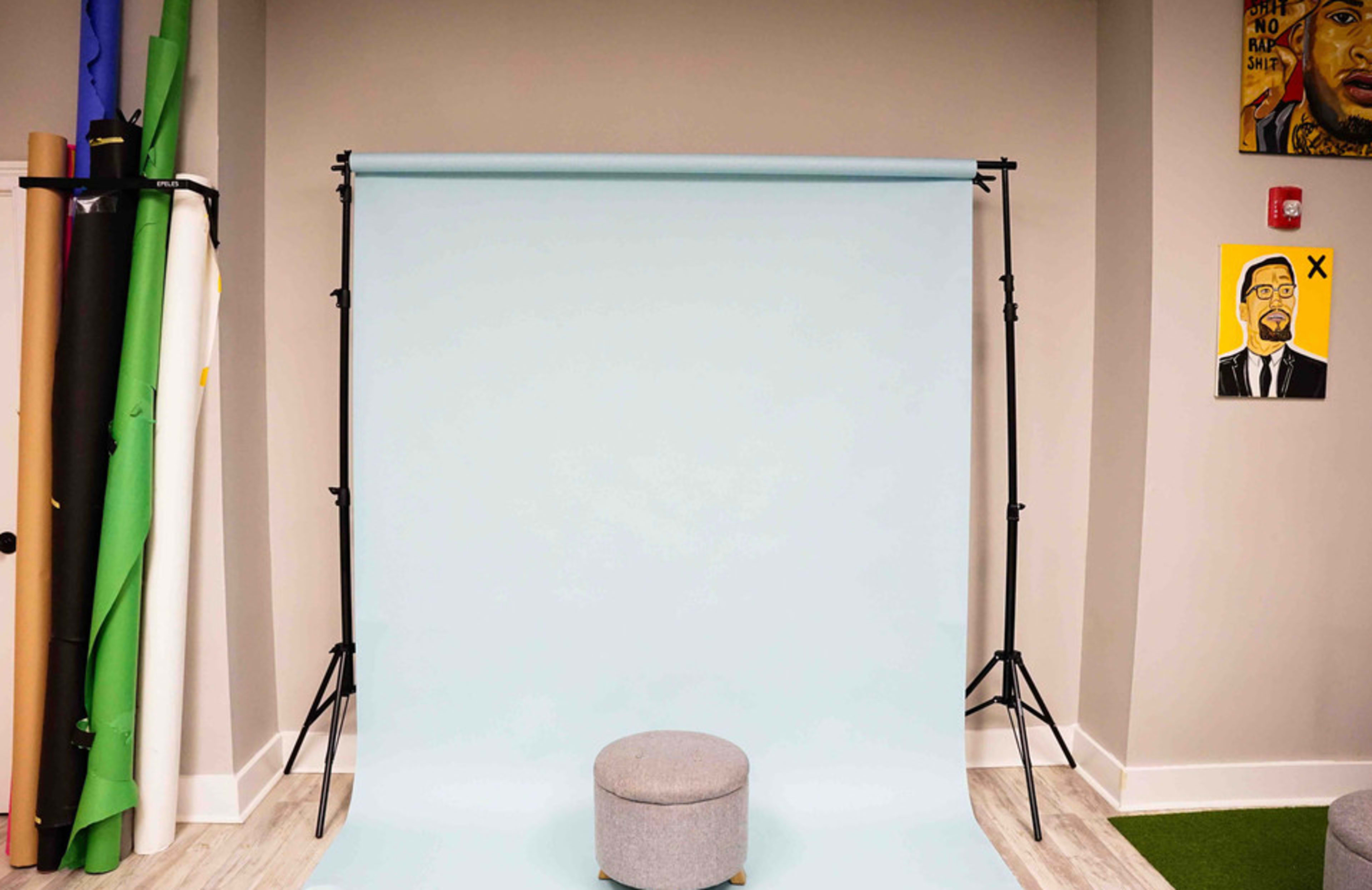 A photography backdrop of light blue fabric with a gray stool in front of it, surrounded by colorful backdrop rolls and framed art on the walls.