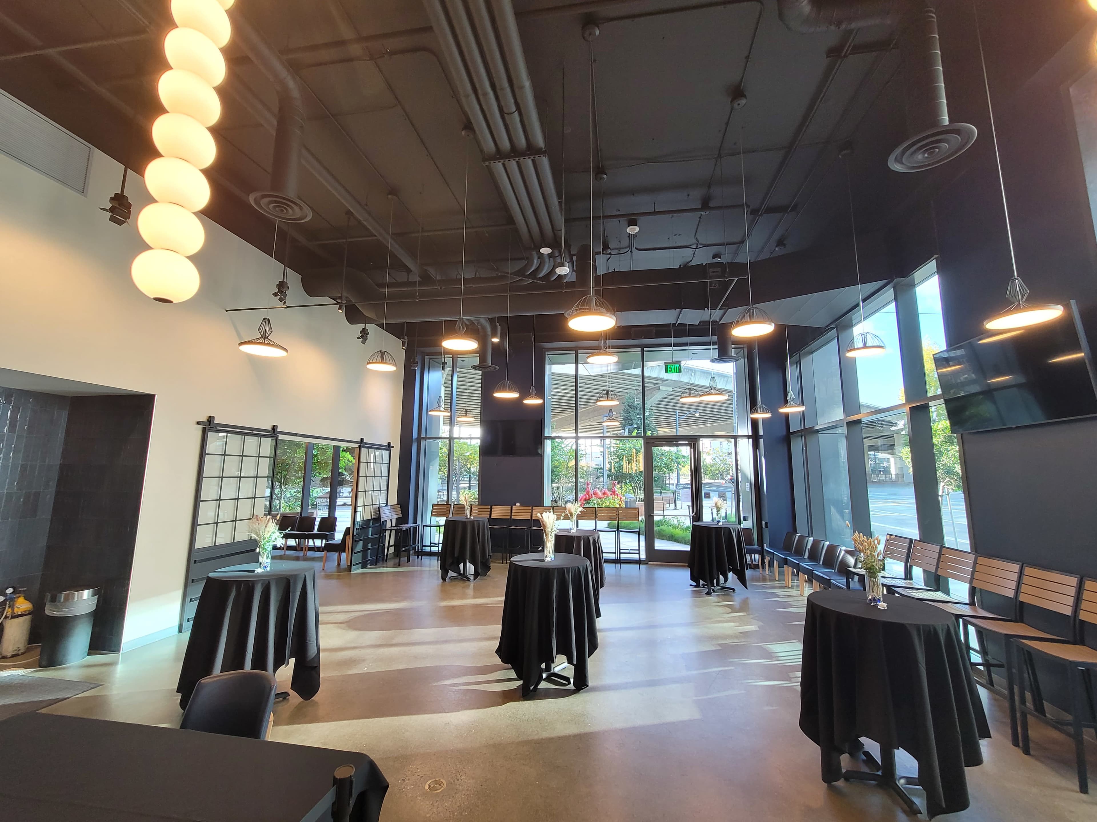 The image shows an empty event space featuring several round tables covered with black tablecloths, modern lighting fixtures, and large windows letting in natural light.