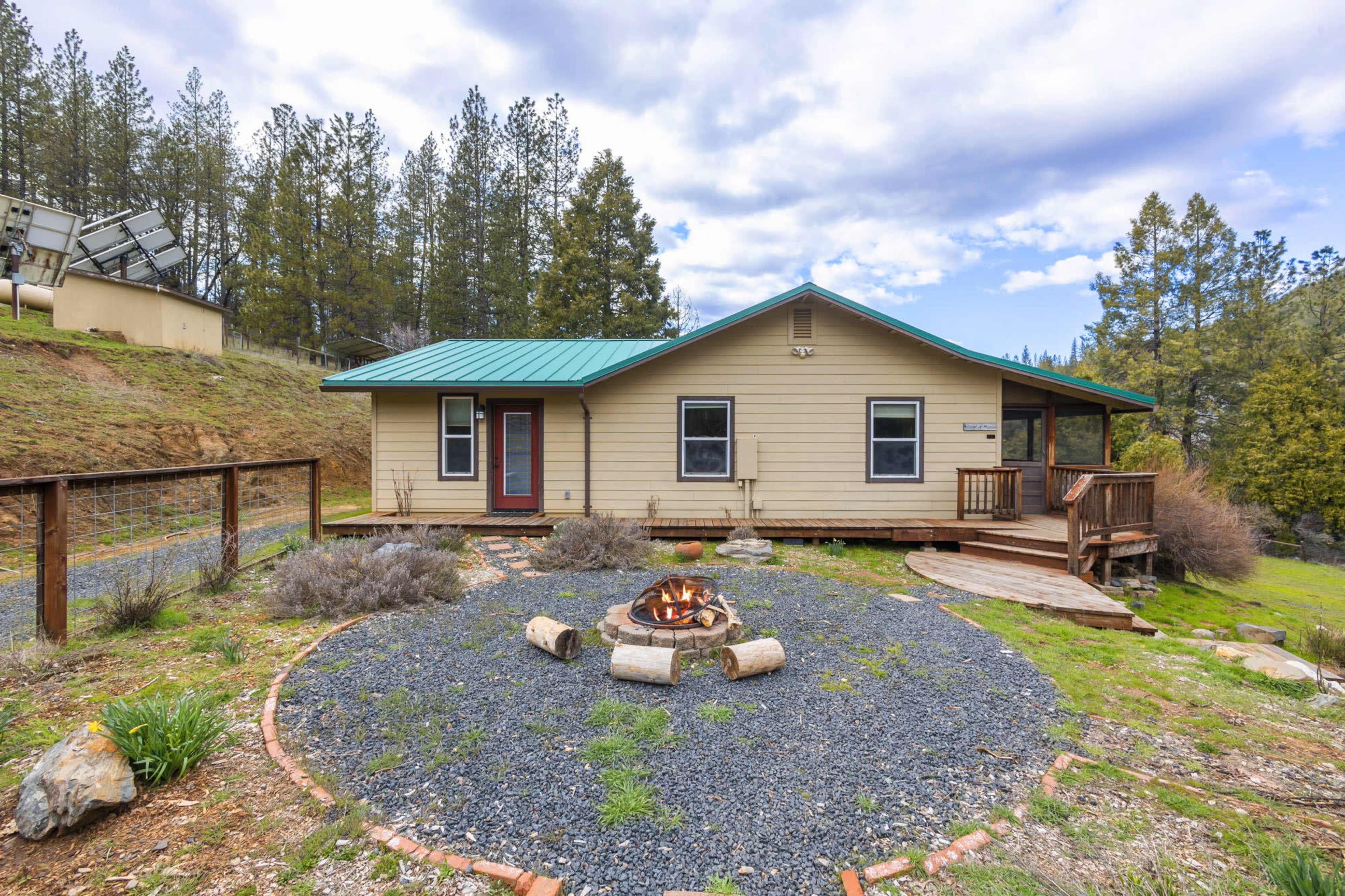 A single-story house with a green metal roof is situated near a circular fire pit surrounded by gravel and landscaping in a wooded area.