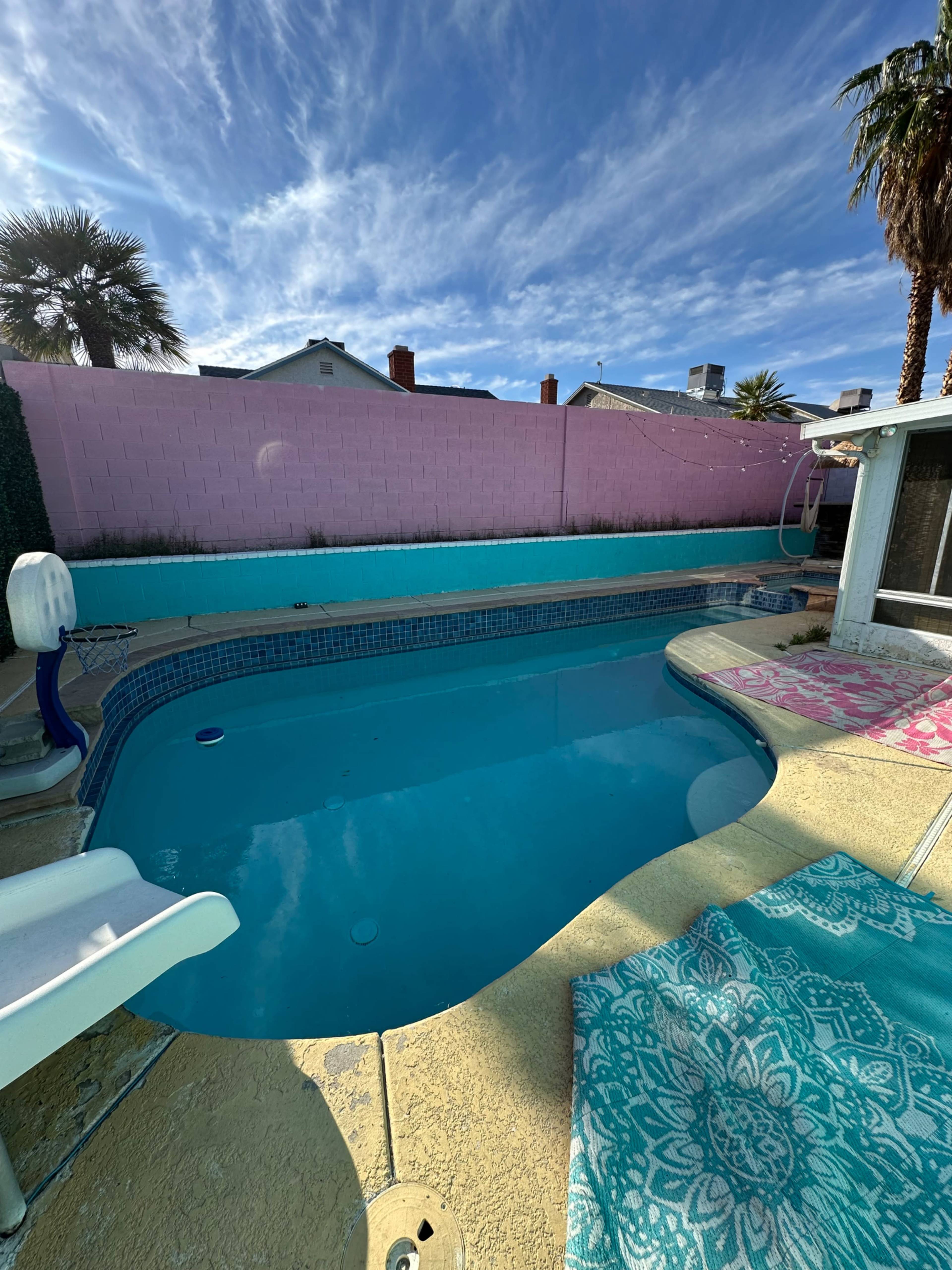 The image shows a swimming pool surrounded by a pink wall and palm trees under a blue sky with scattered clouds.