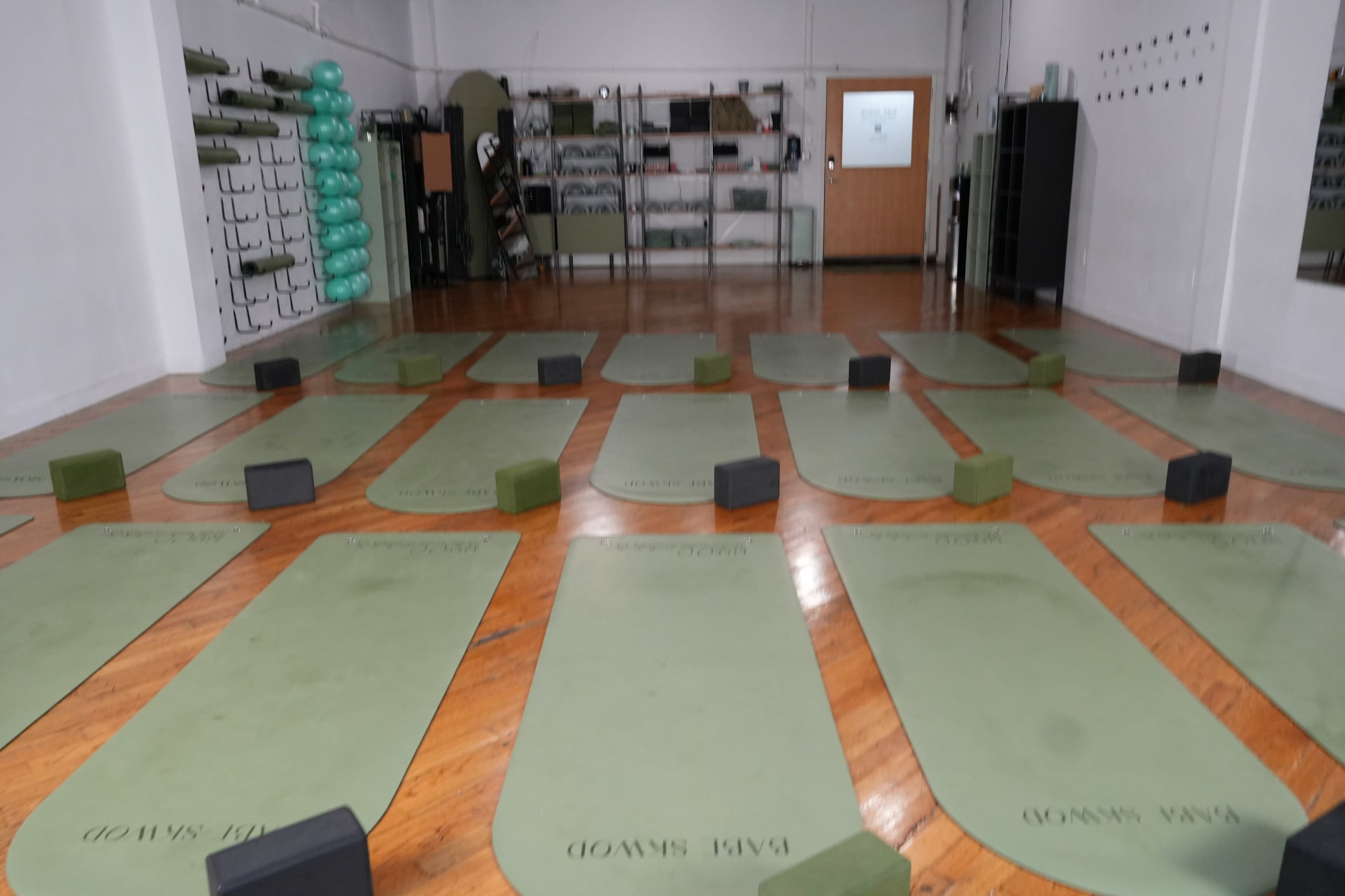 The image shows a spacious yoga studio with green mats arranged in rows, complemented by yoga blocks and wall-mounted storage for exercise equipment.