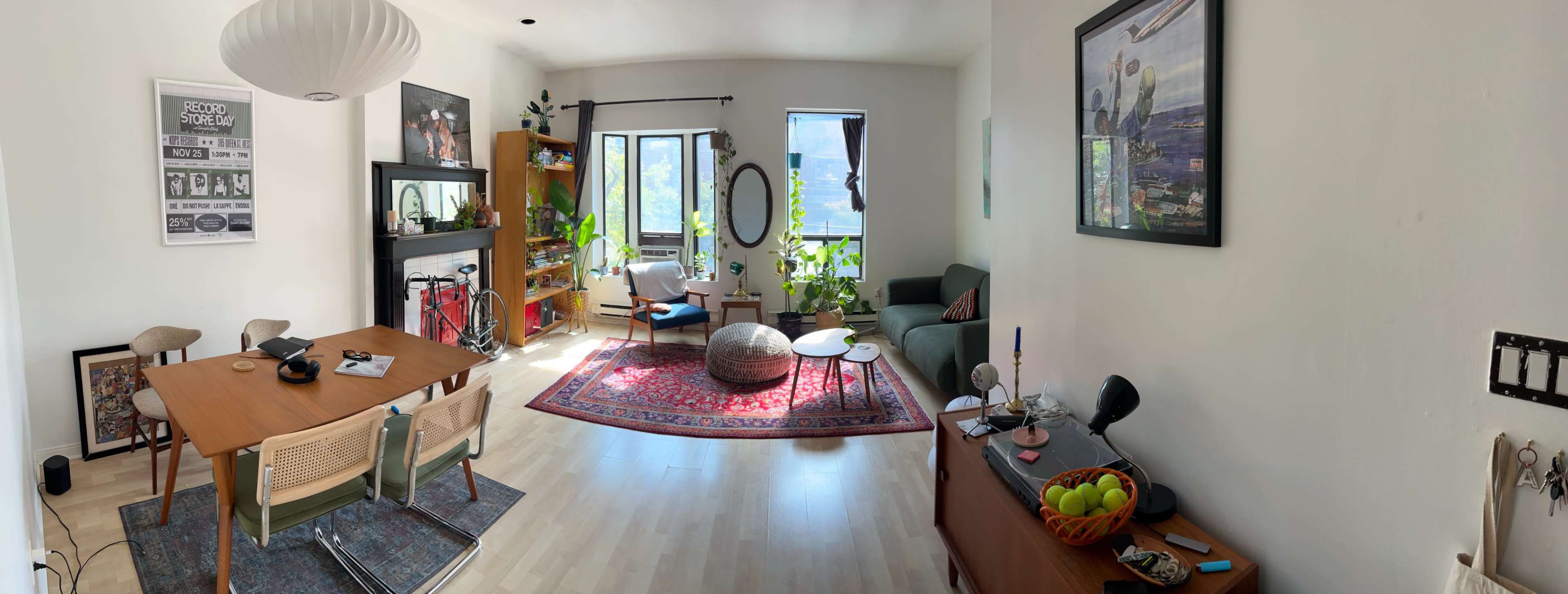 The image shows a cozy living room with a dining table, plants, and a mix of seating arrangements, featuring a large window that lets in natural light.