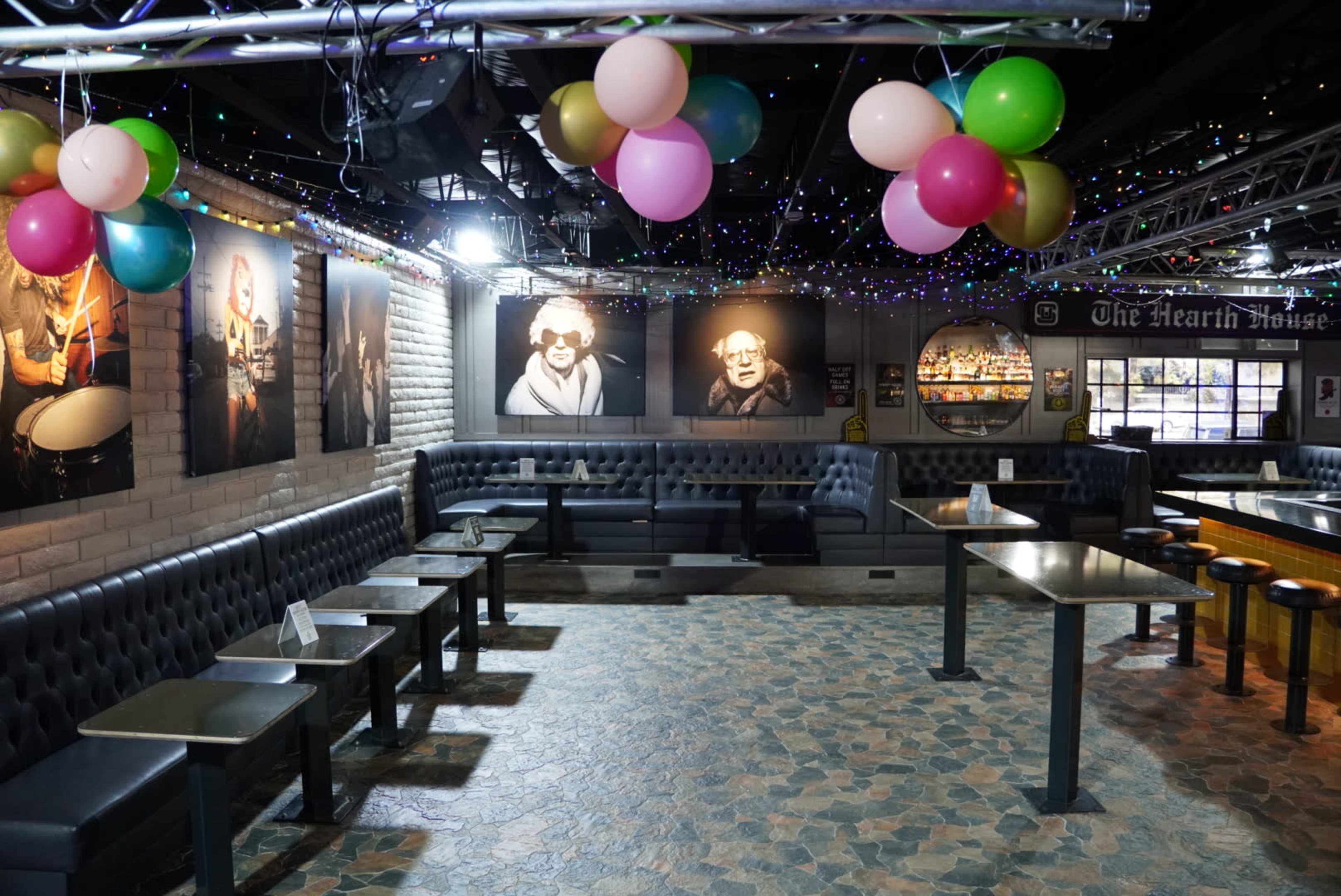 The image shows a dimly lit venue featuring leather seating, tables, and colorful balloons suspended from the ceiling, with black-and-white photographs lining the walls.