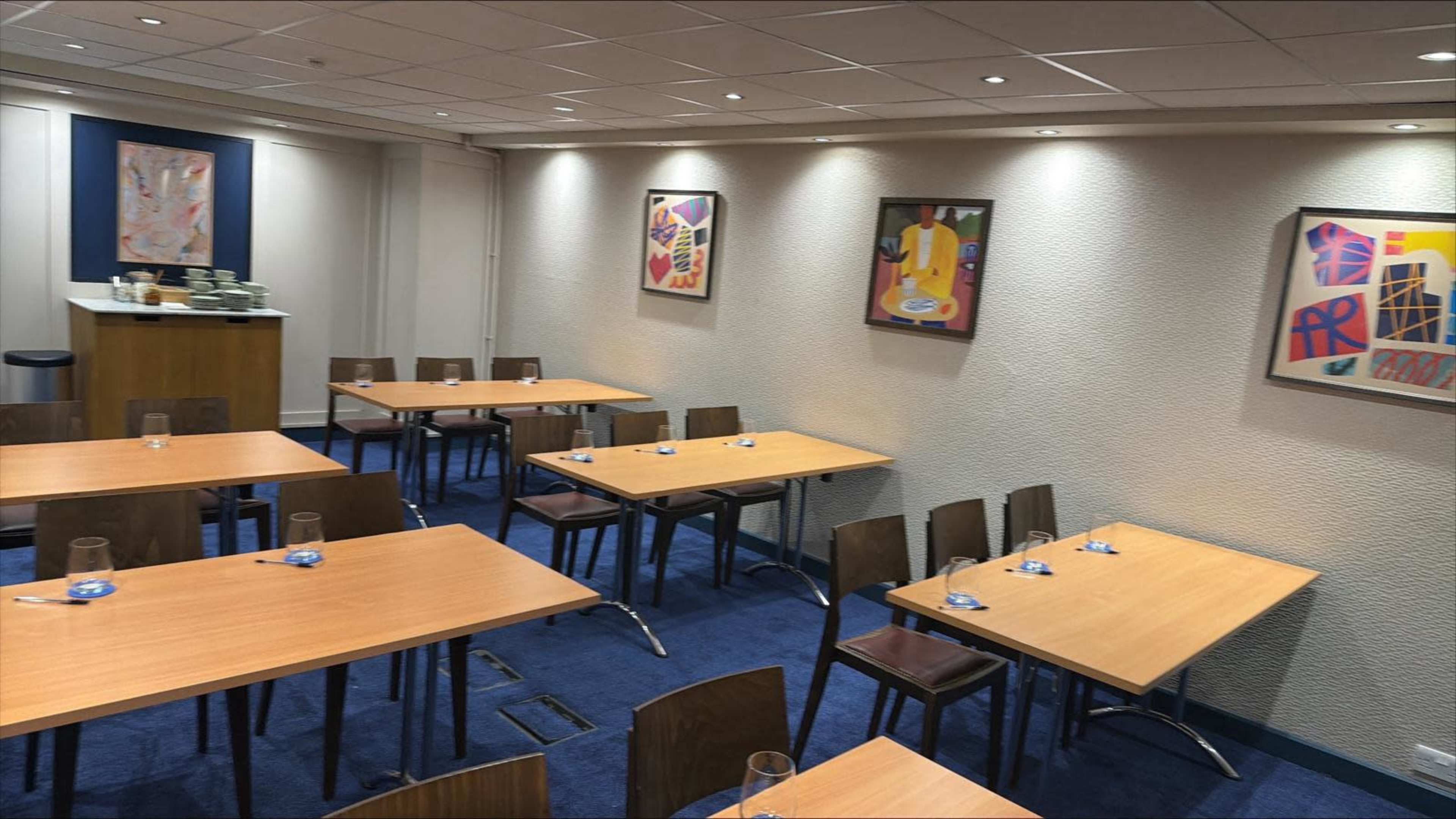 The image shows a restaurant or meeting room with several wooden tables arranged in a grid pattern, surrounded by chairs, and artwork displayed on the walls.