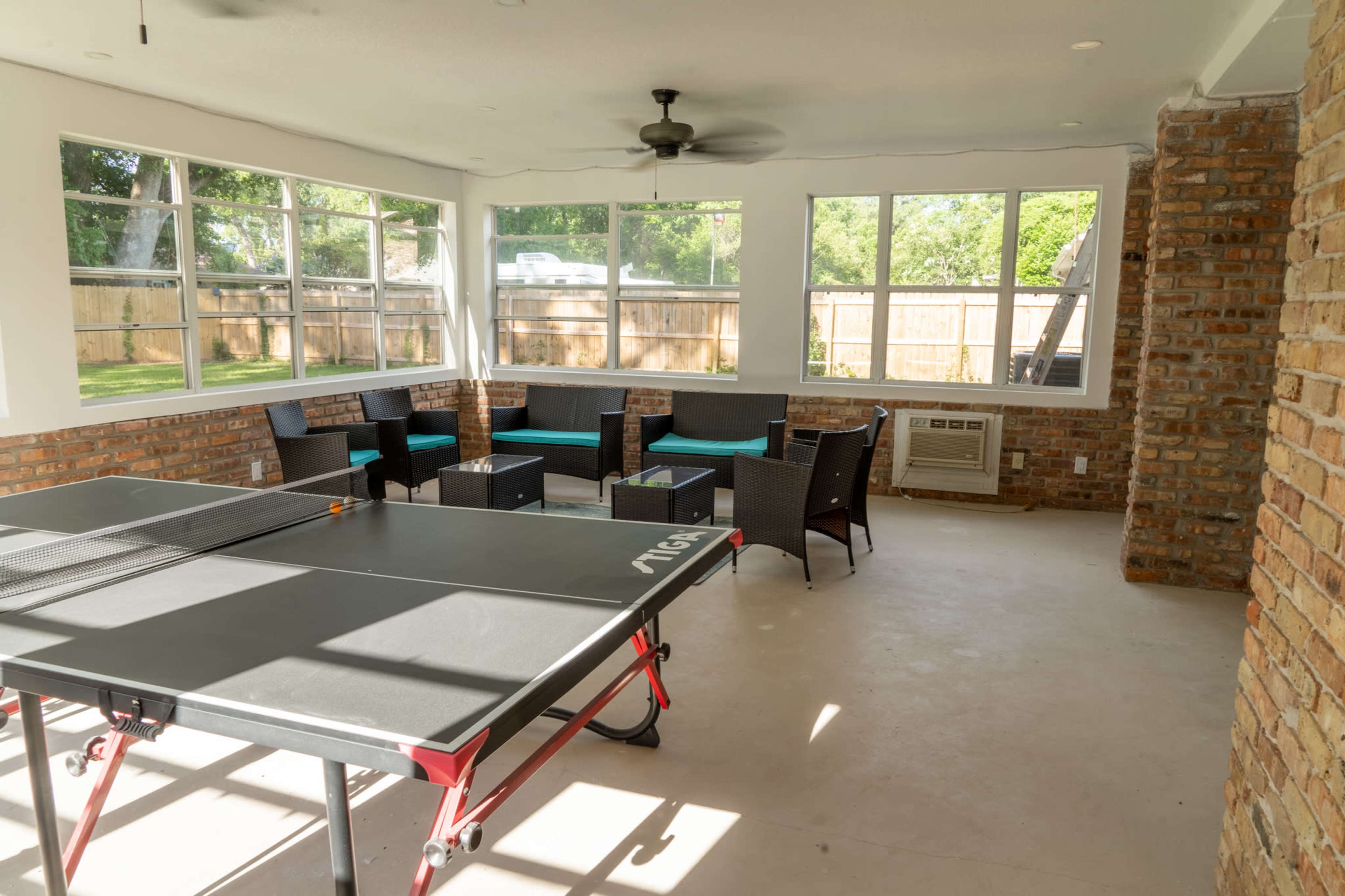 A ping pong table is placed in a sunlit room with large windows, surrounded by seating and brick walls.