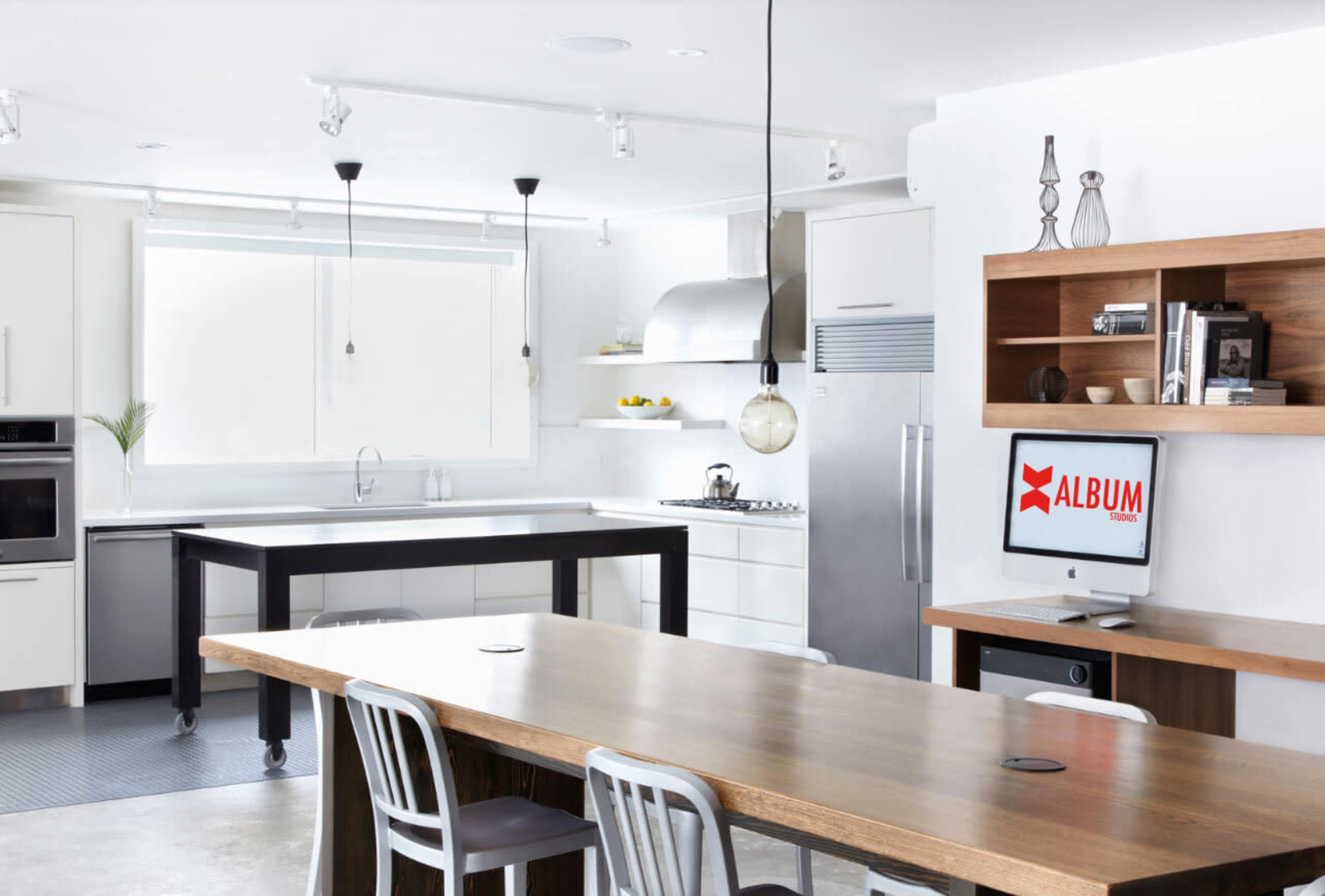A modern kitchen with a large wooden dining table, sleek appliances, and a computer on a desk in the corner.