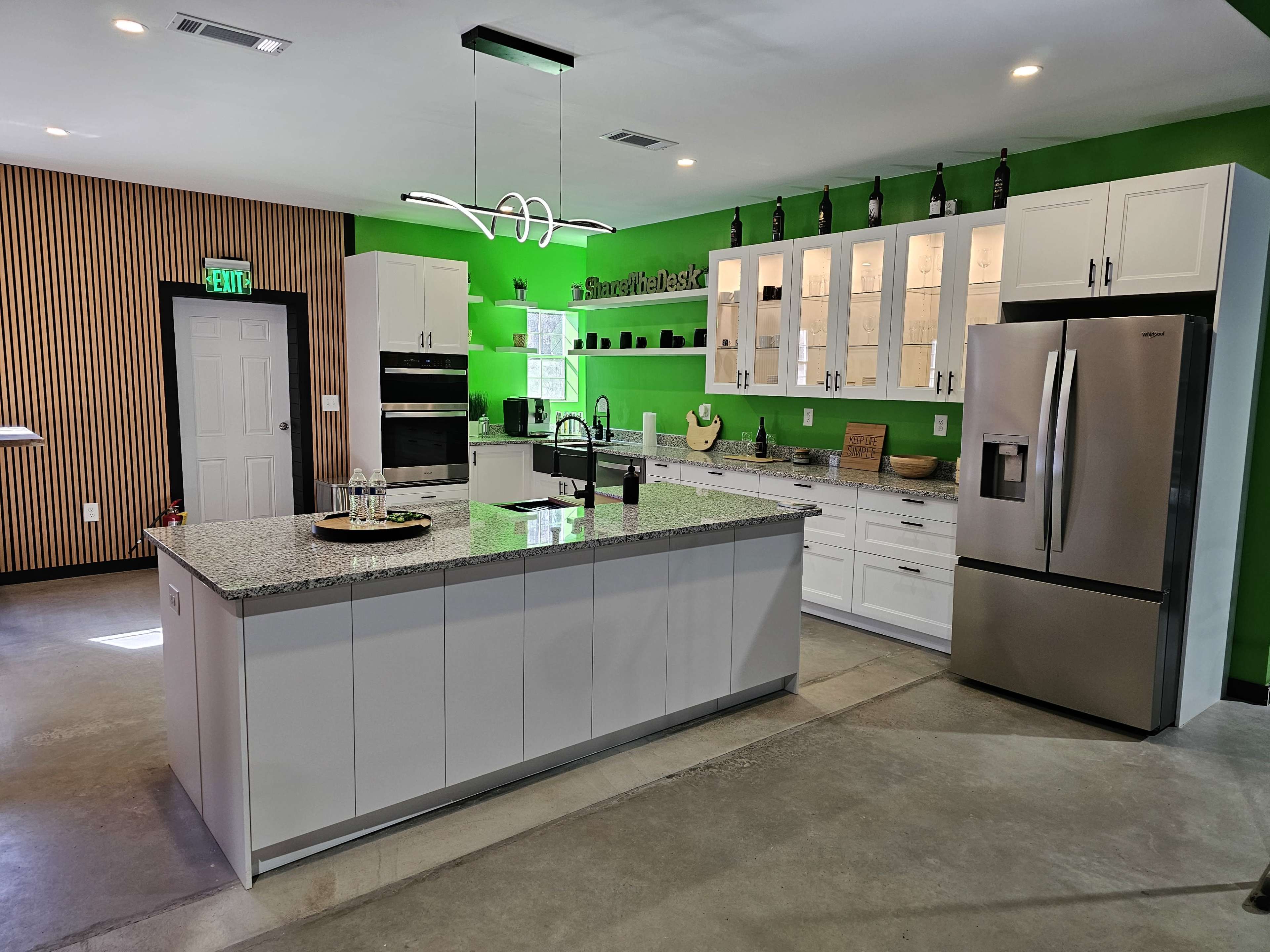 The image shows a modern kitchen with a central island, granite countertops, white cabinetry, and a green accent wall adorned with wine bottles.