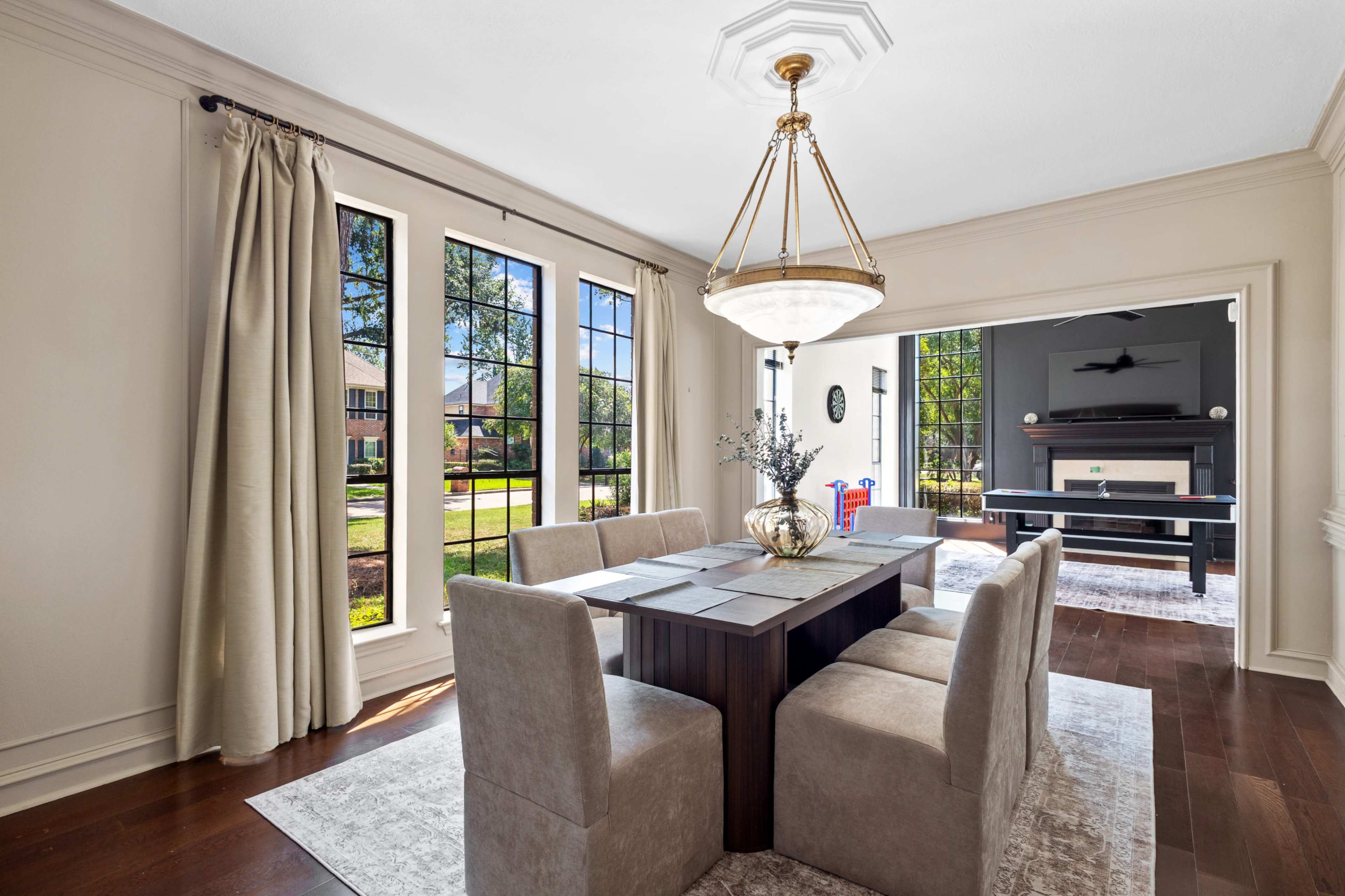 A dining room features a long wooden table surrounded by six upholstered chairs, large windows allowing natural light, and an open view to an outdoor space.