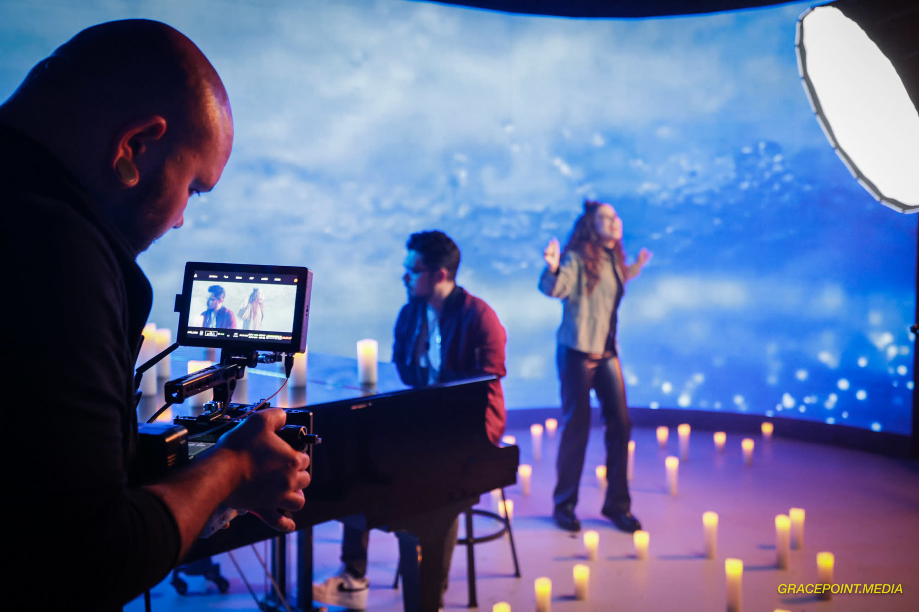 A musician performs in front of a blue backdrop while a camera operator captures the scene, surrounded by lit candles.