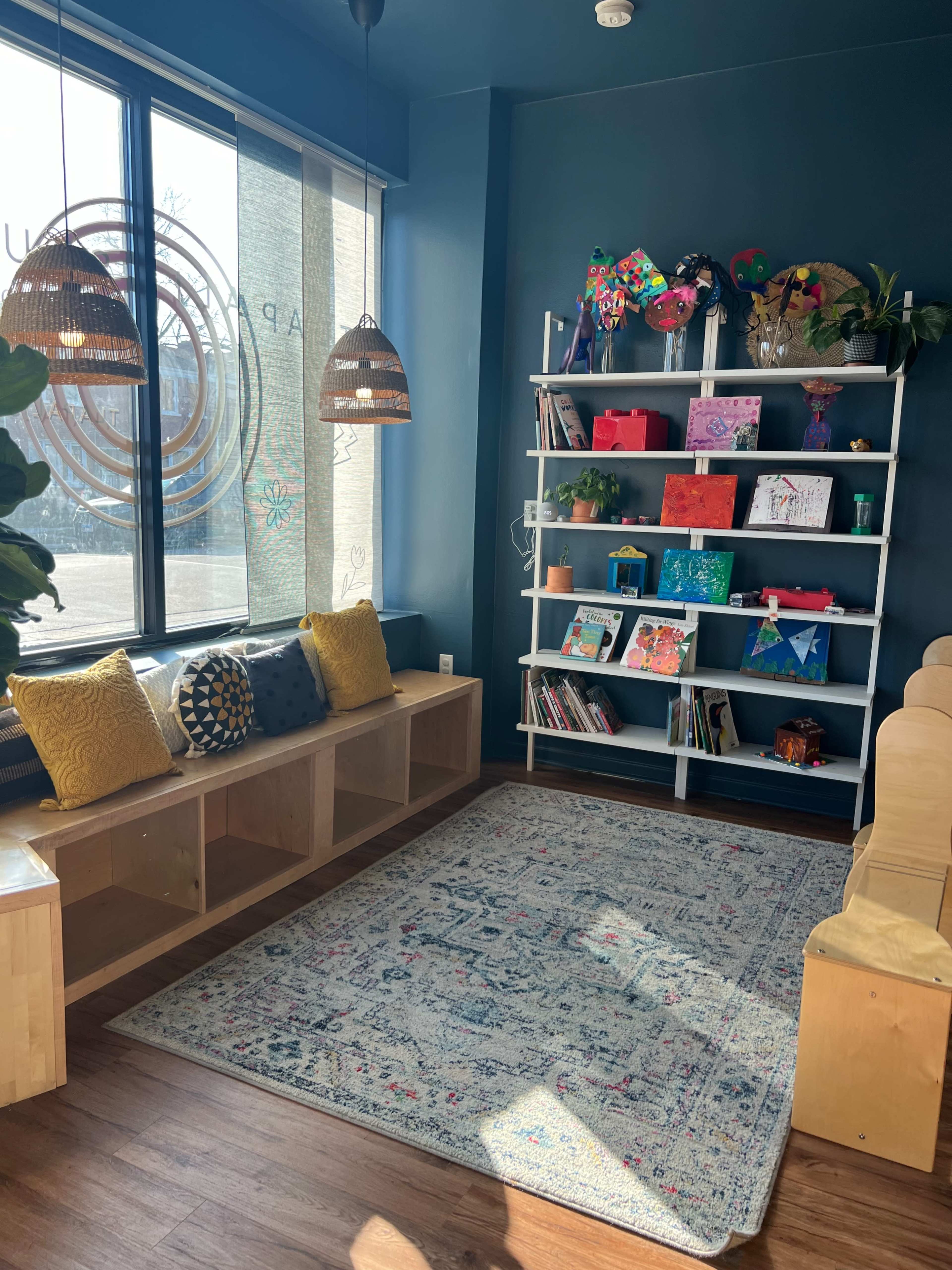The image shows a calm, cozy reading nook with a blue wall, shelves filled with colorful books and art, a patterned rug, and a bench with cushions.