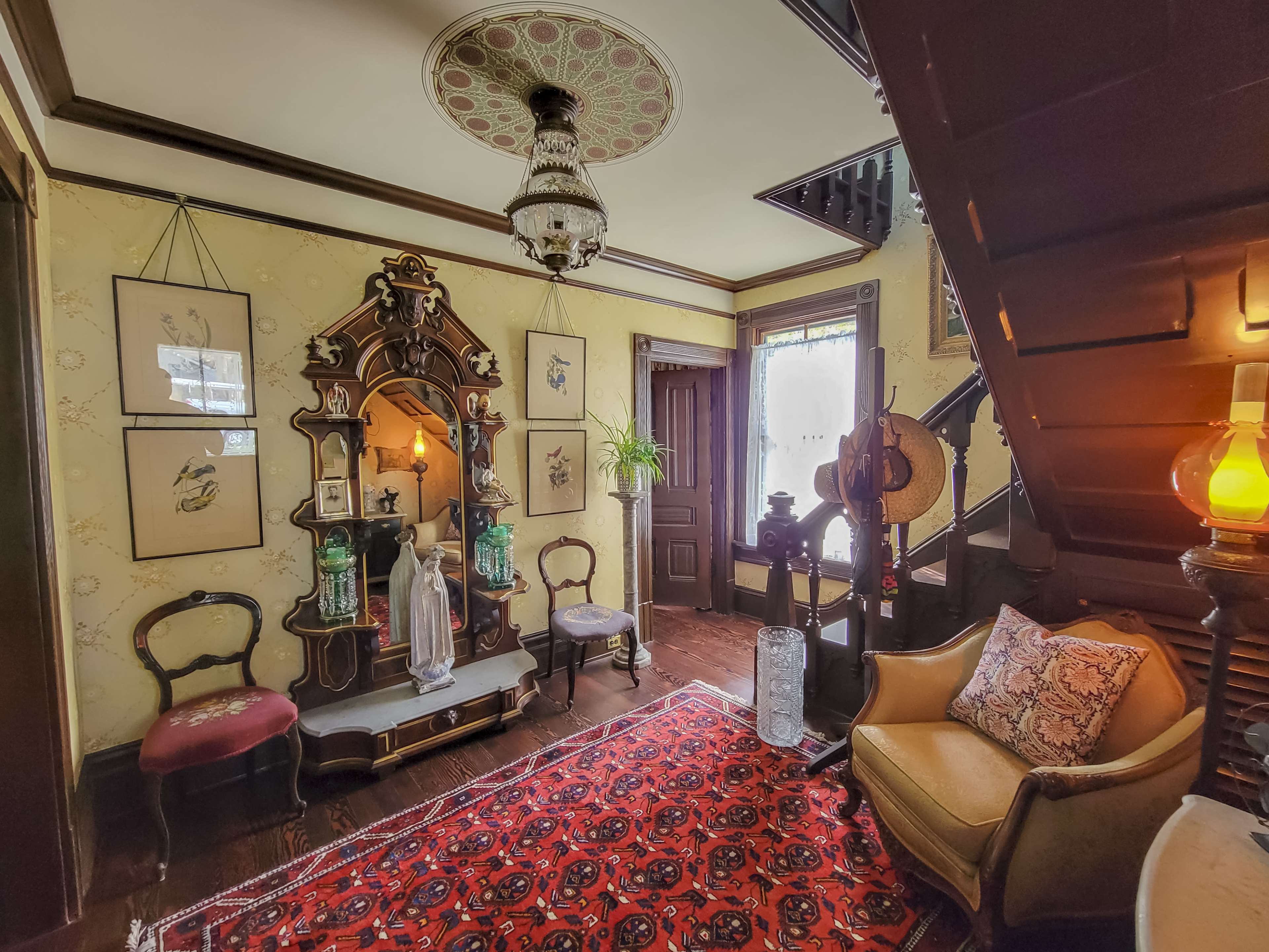 The image shows a vintage interior with ornate wooden furniture, framed artwork on the walls, and a staircase leading to an upper level.