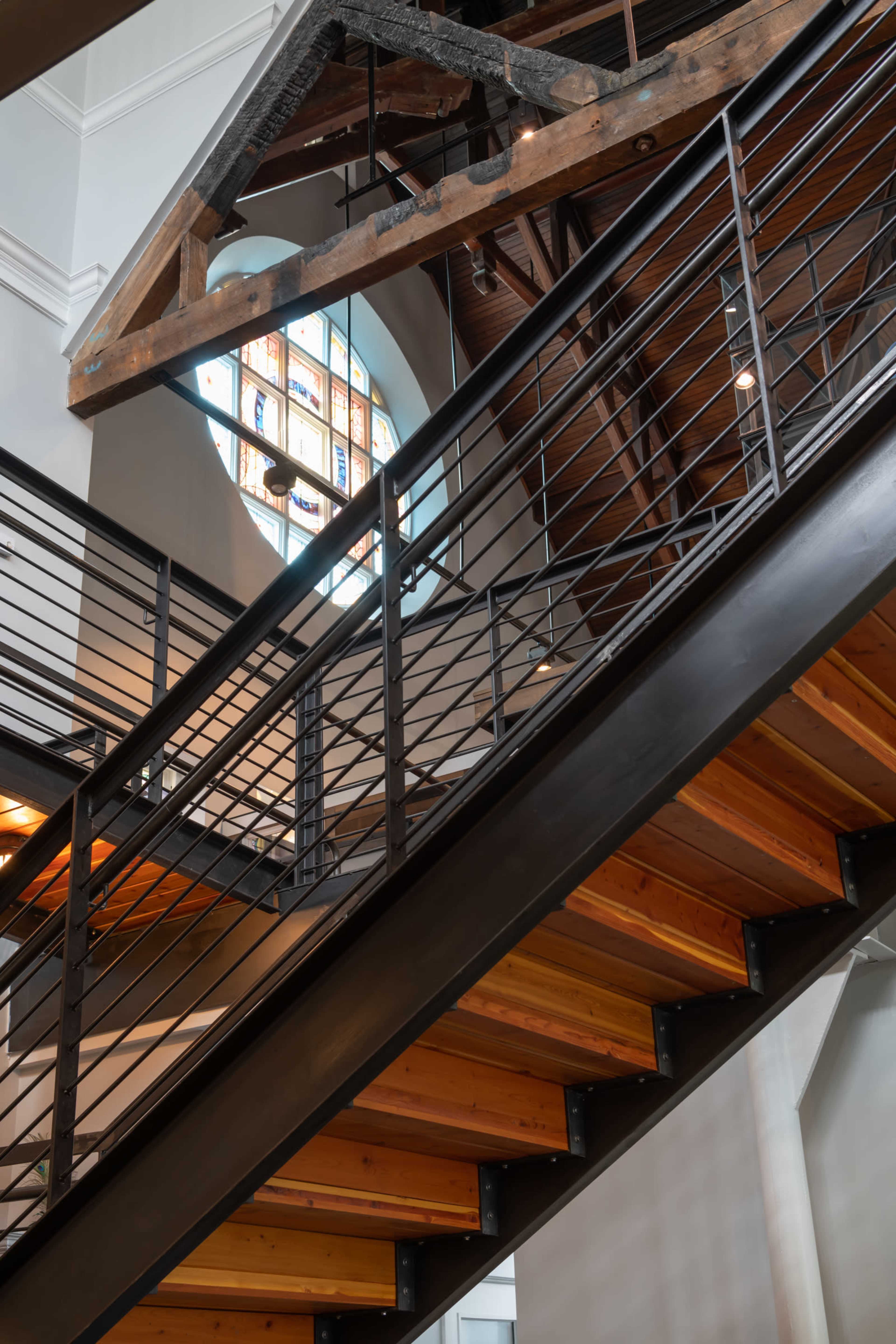 A staircase with wooden steps and a metal railing, leading to a circular stained glass window in a building with exposed wooden beams.