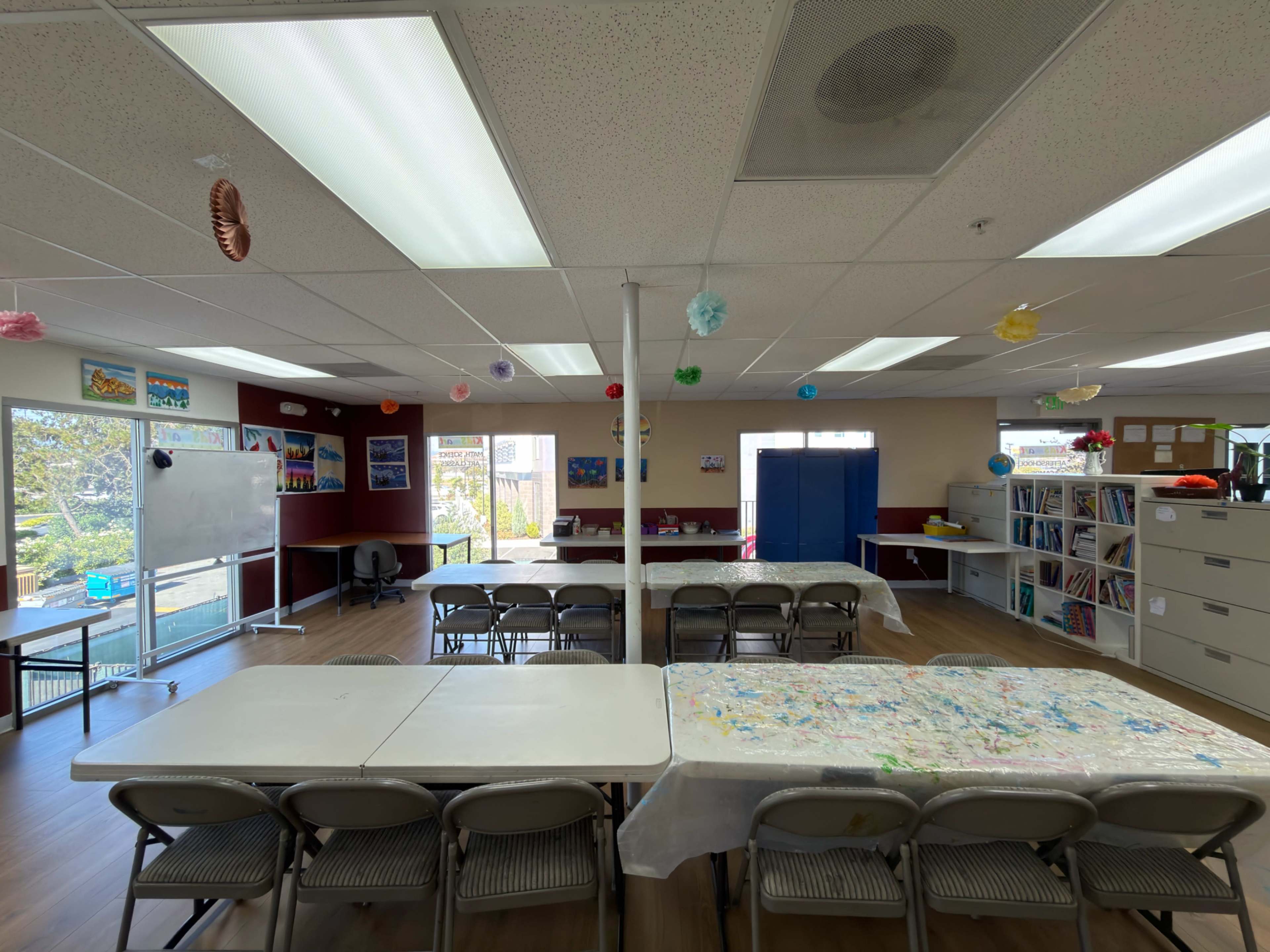 A brightly lit classroom features several tables covered with white tablecloths and is decorated with colorful hanging ornaments.
