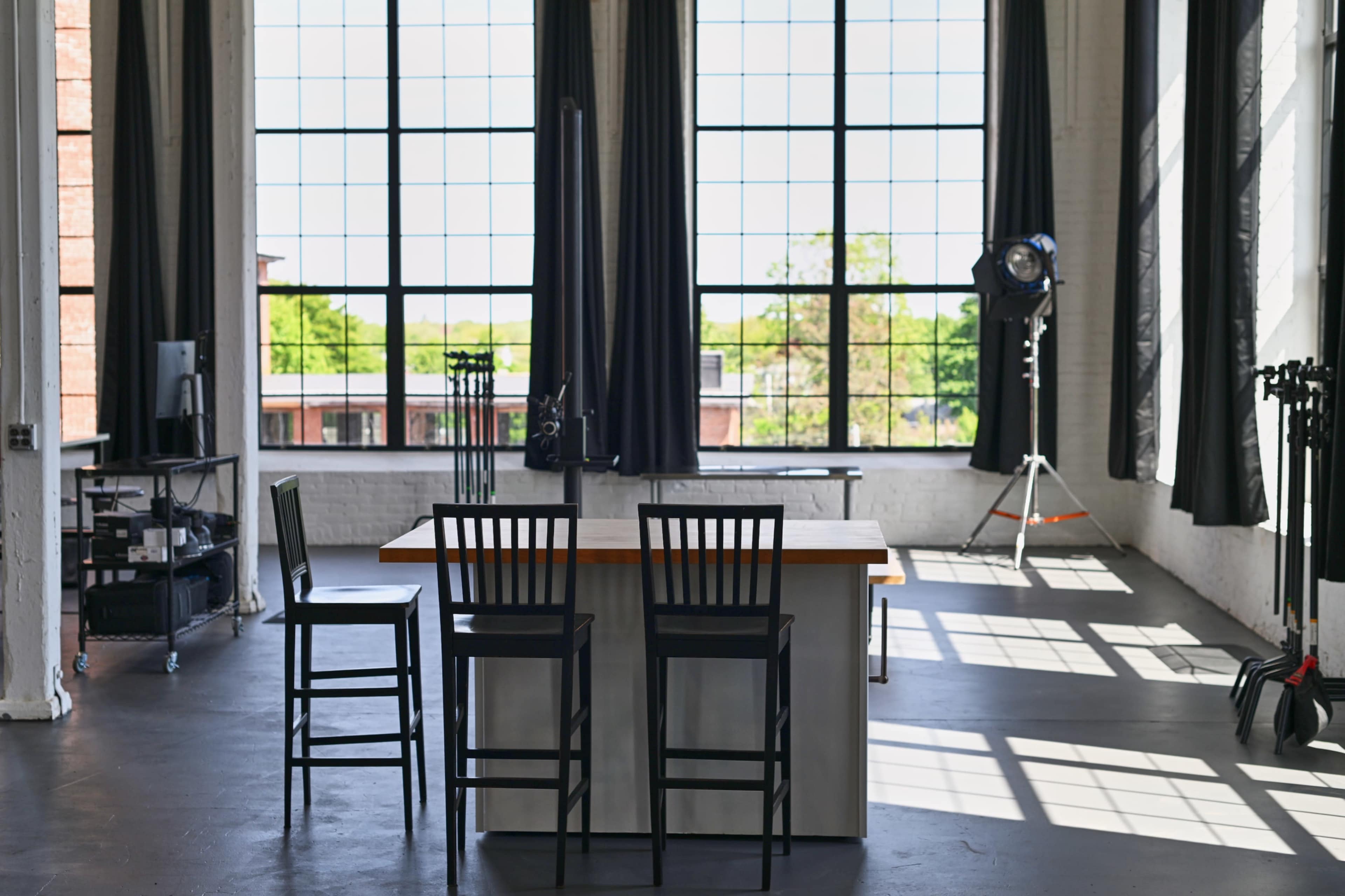 A modern studio space features a central island with three black bar stools, large windows allowing natural light, and various equipment in the background.