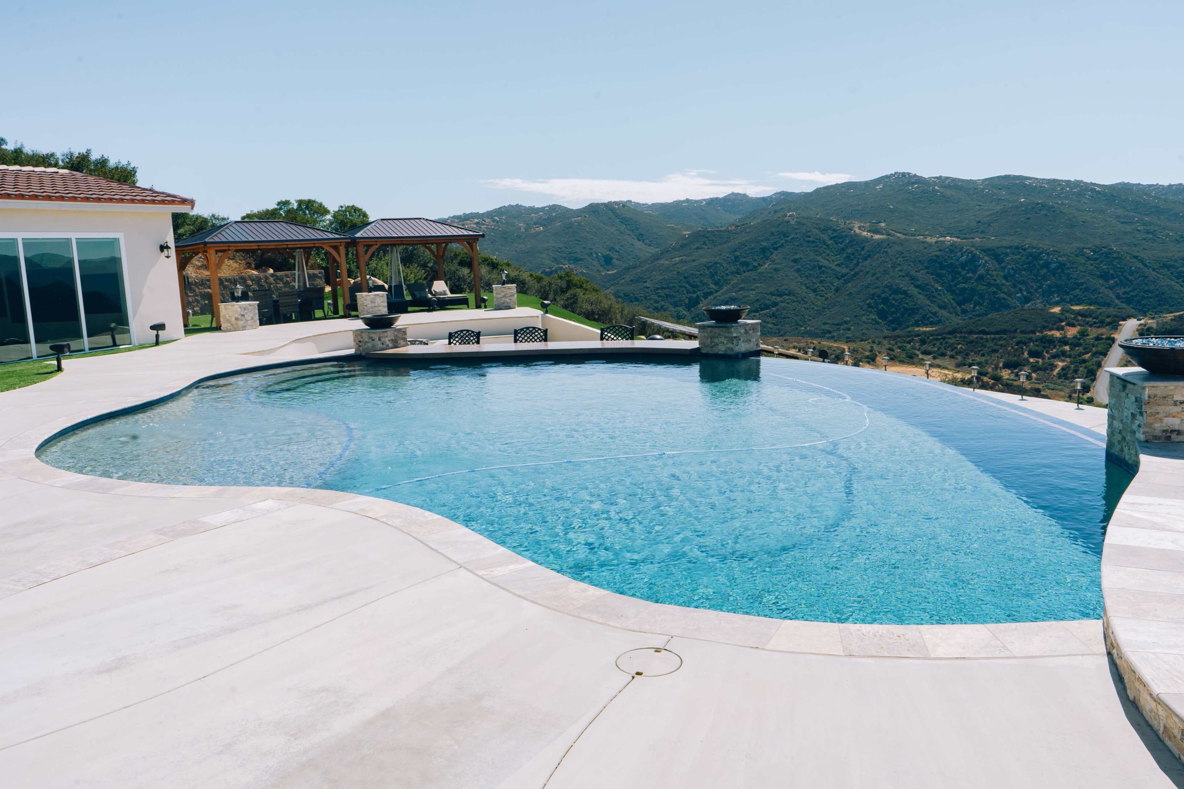 A modern pool overlooking a mountainous landscape, with a patio and seating areas nearby.