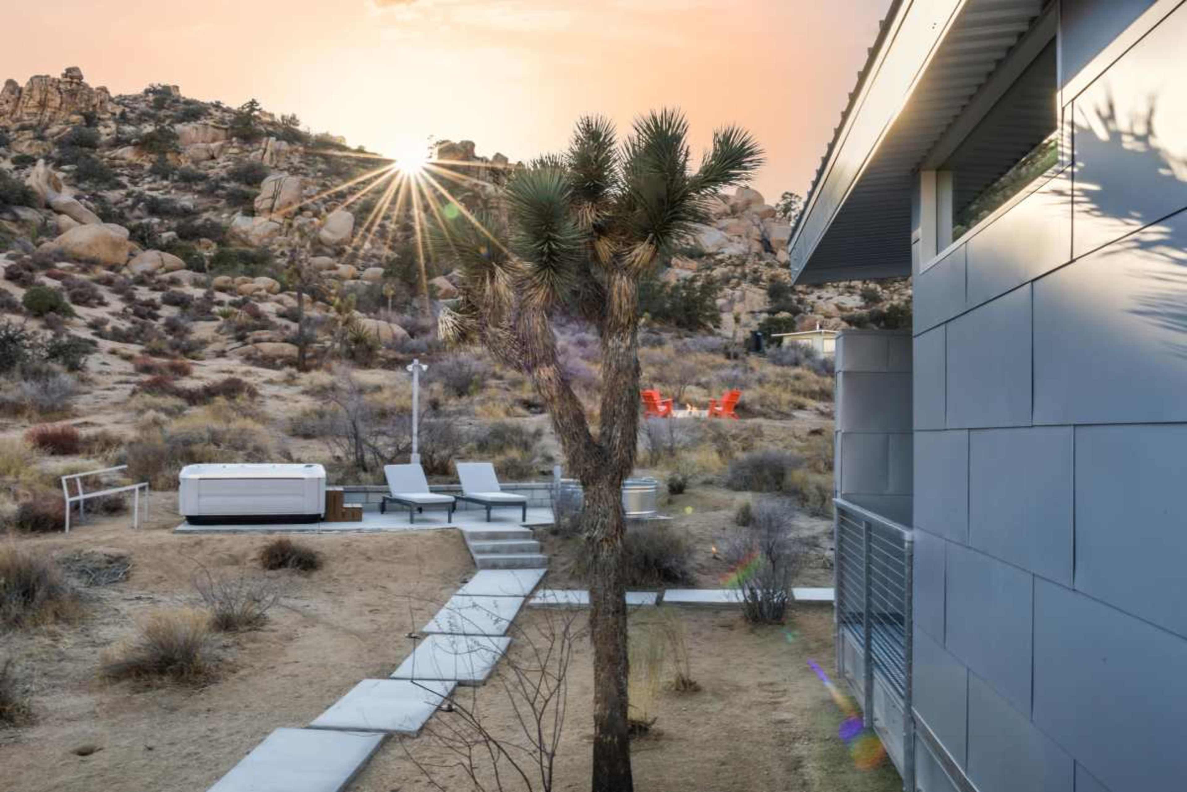 The image shows a modern structure next to a pathway leading to a hot tub, with a Joshua tree and rocky hills in the background as the sun sets.