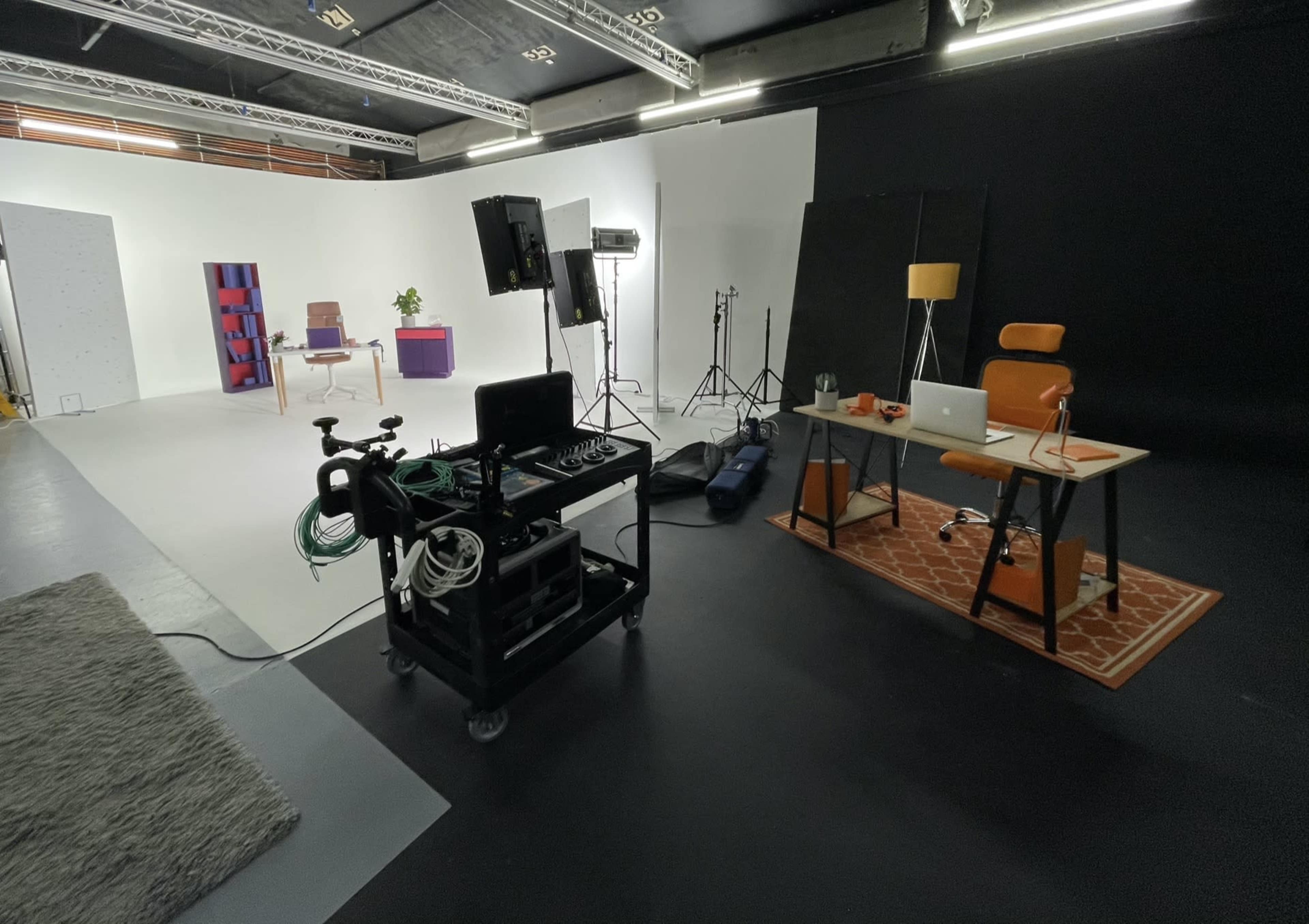 A studio setup with a desk, chair, and equipment on carts, alongside various lighting and backdrop elements in an enclosed space.