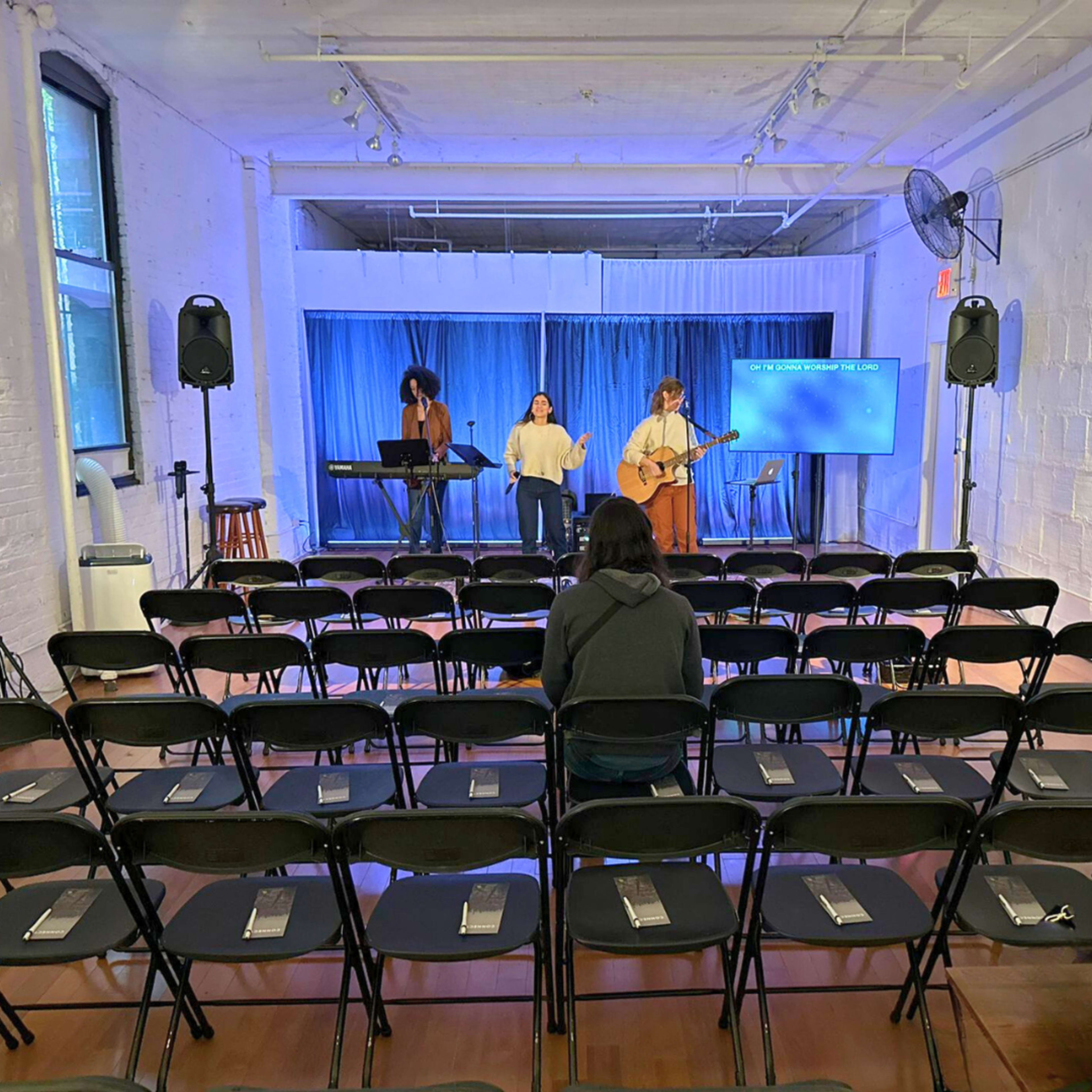 Three musicians are performing on a stage in a nearly empty room filled with black folding chairs facing the performers.