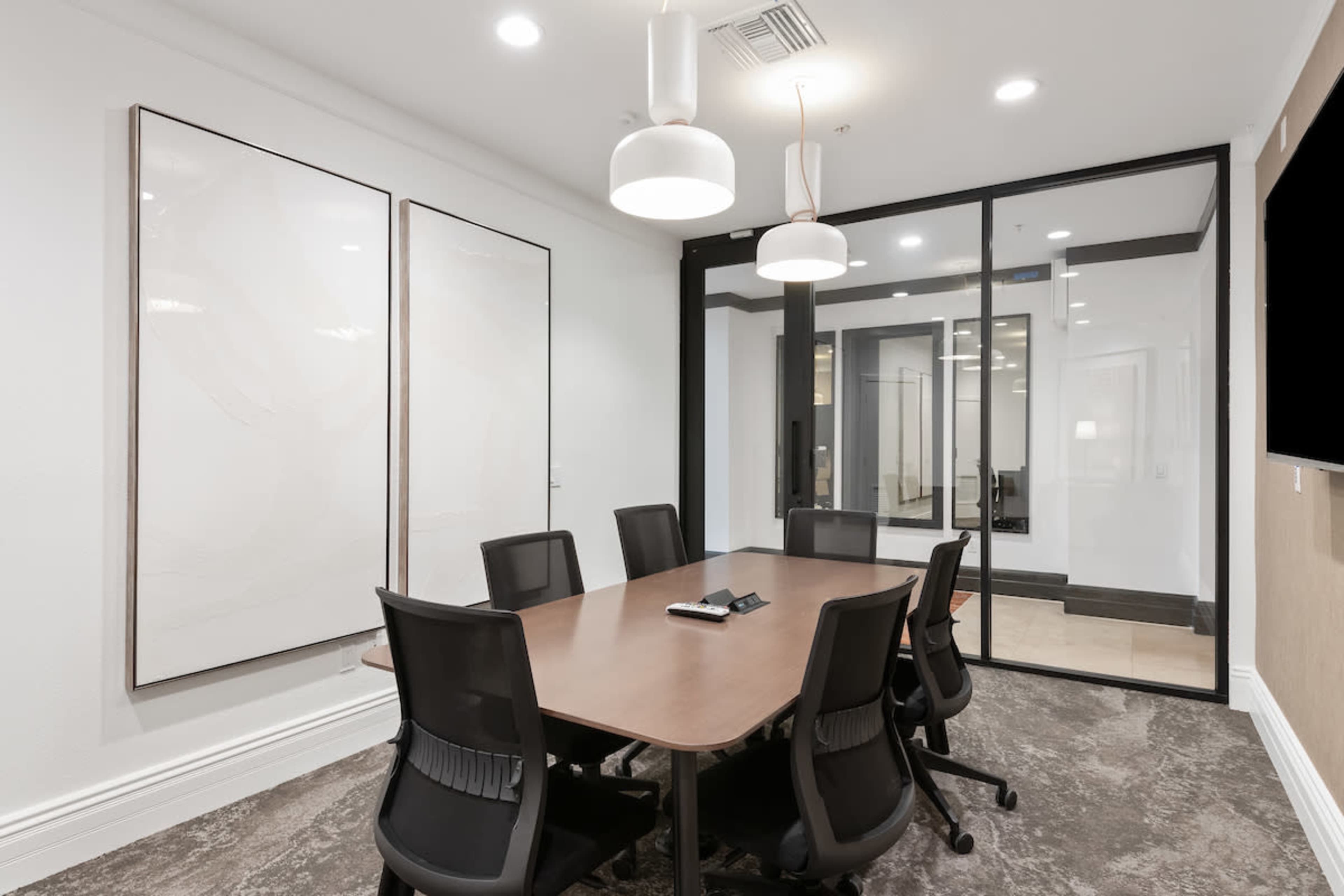 A modern conference room features a large table surrounded by black chairs, with two blank framed artworks on the walls and glass partitions separating it from an adjacent area.