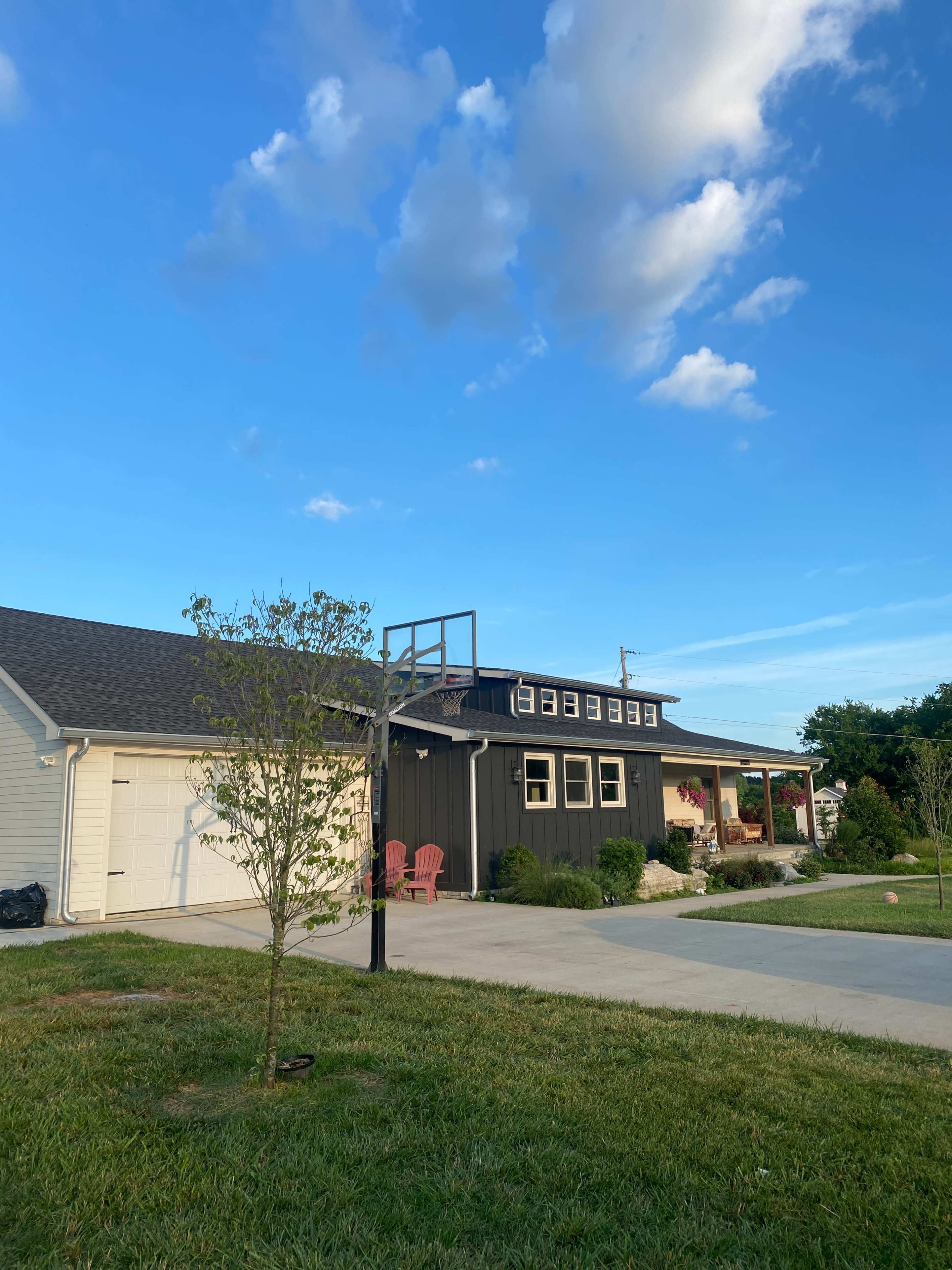 A two-story black and white house with a basketball hoop stands next to a driveway lined with green grass under a blue sky with scattered clouds.