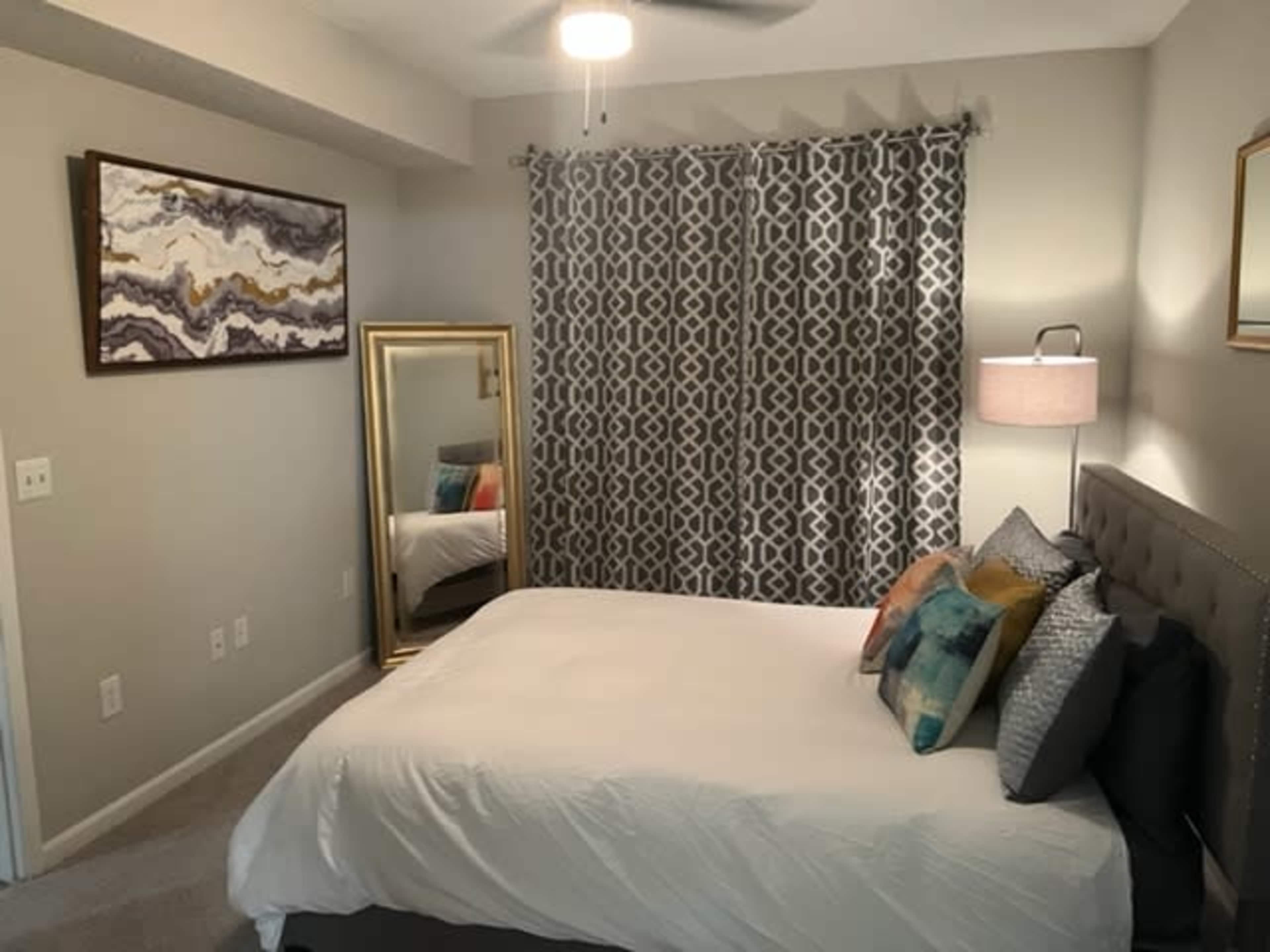 The image shows a neatly arranged bedroom with a large bed, decorative pillows, a mirror, a lamp, and patterned curtains.