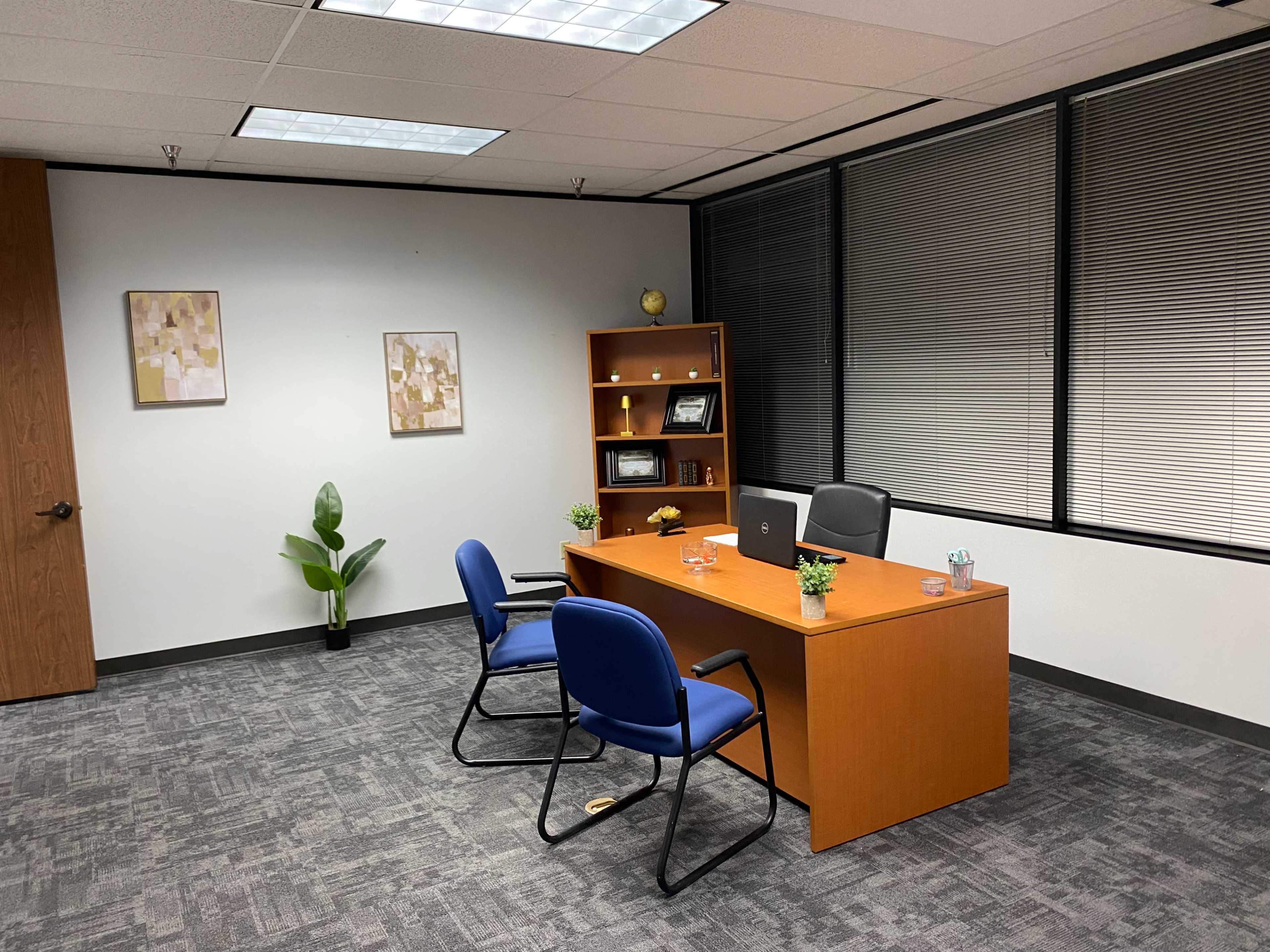 An office space features a wooden desk with two blue chairs, a bookshelf, and a small plant, all set against large windows with blinds.