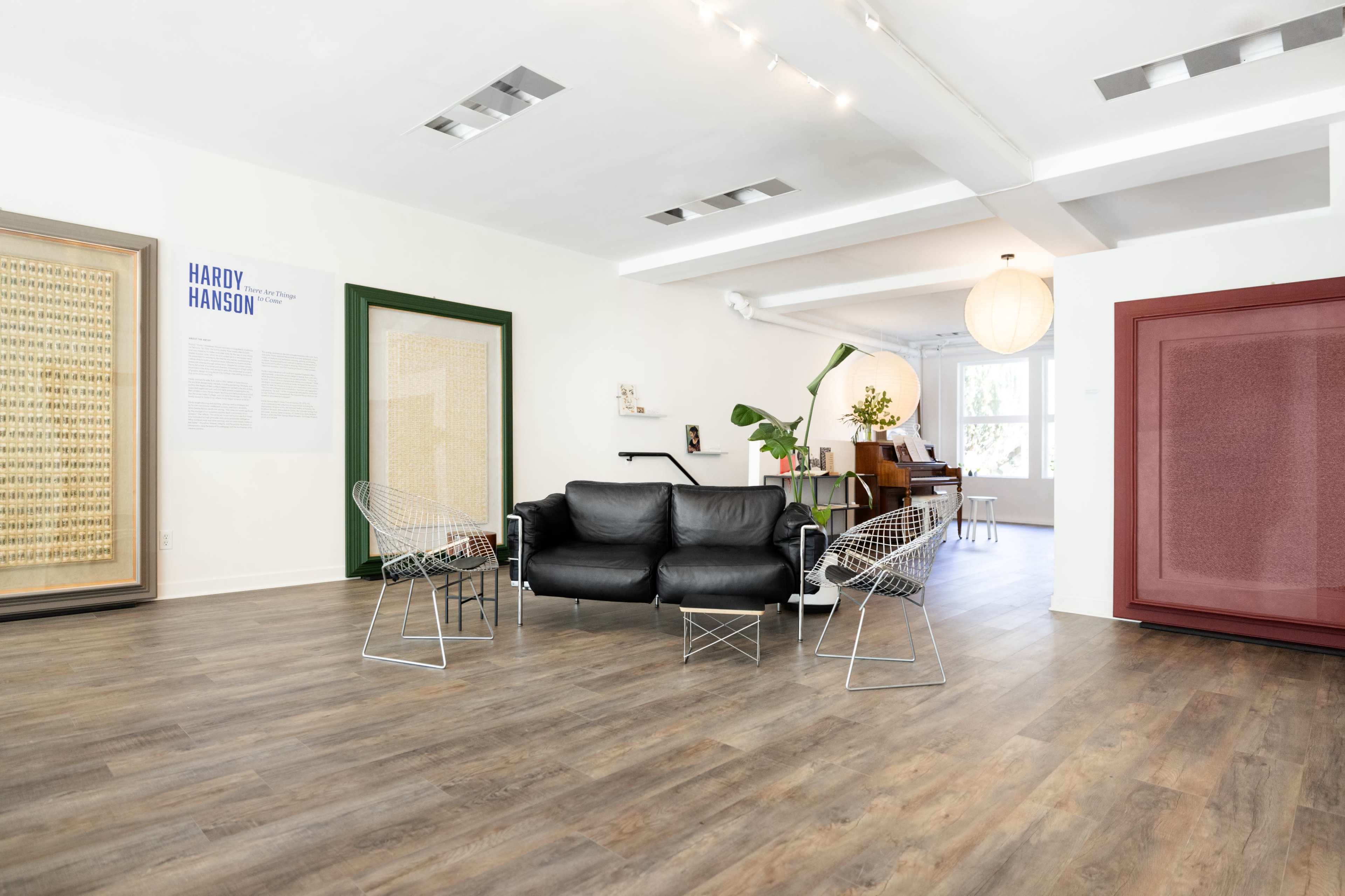 The image shows a modern interior space featuring a black leather sofa, two wire chairs, and colorful framed artwork on the walls.