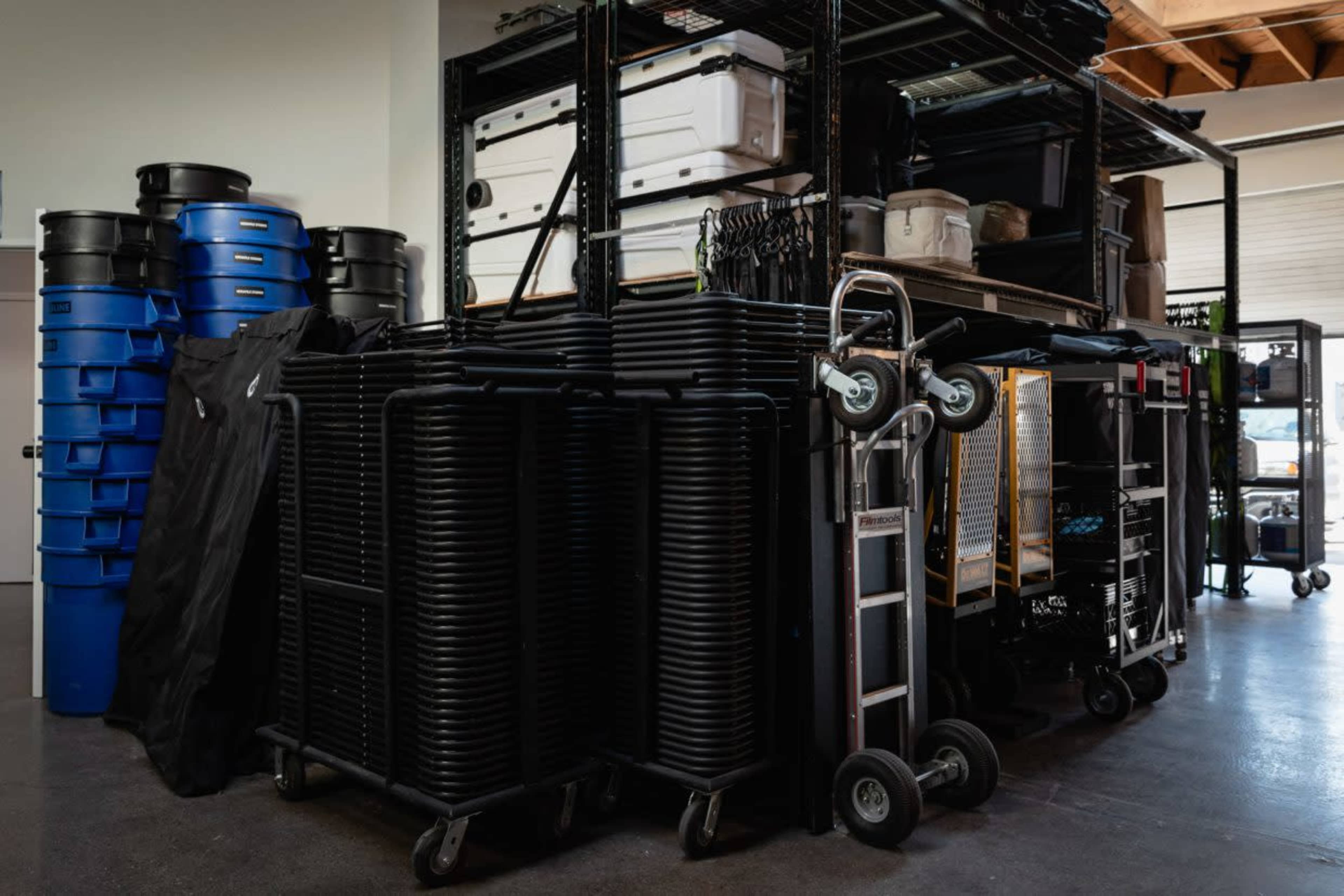 The image shows a storage area filled with stacked black folding chairs, blue bins, and metal shelving units.