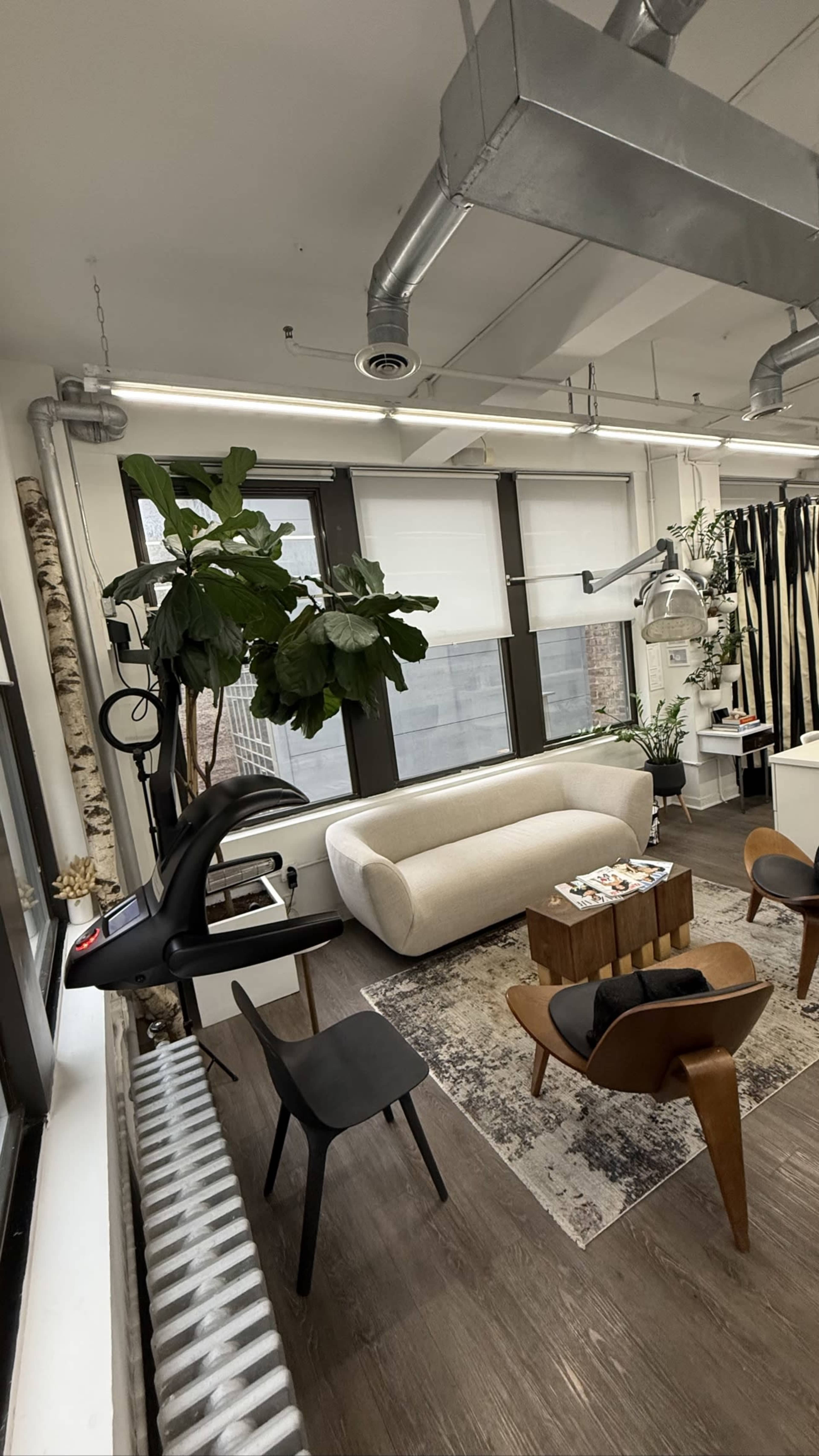 The image shows a modern office space with a beige couch, wooden table, chairs, and plants near large windows.