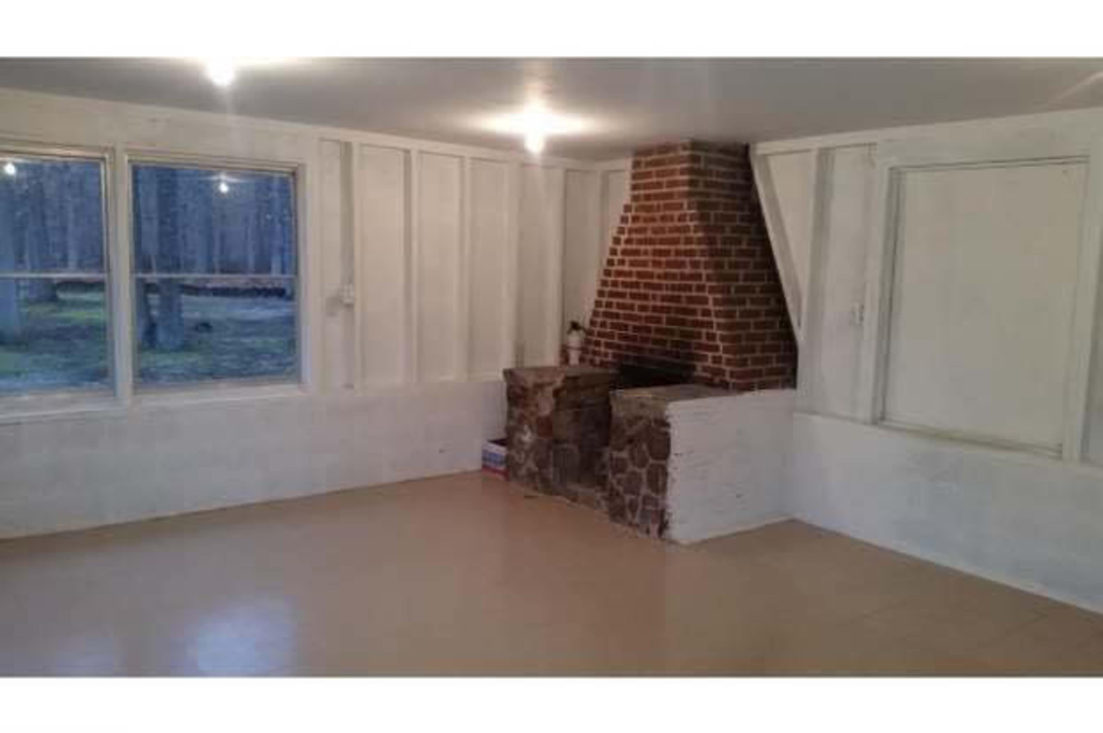 The image shows a spacious room with wooden walls, a brick fireplace, and large windows facing a wooded area.