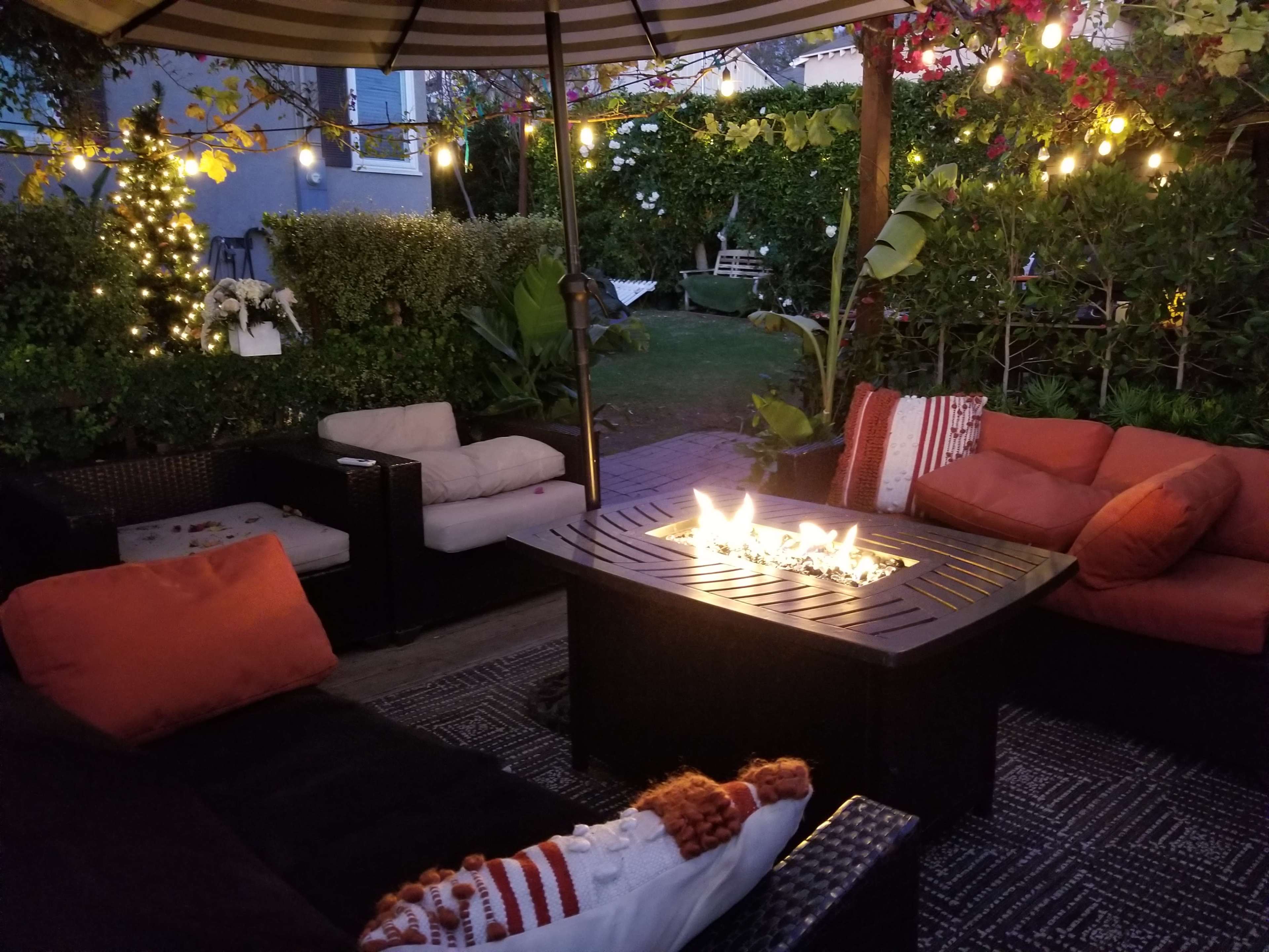 A cozy outdoor seating area features a fire pit surrounded by couches and decorative pillows, with string lights illuminating the lush greenery in the background.