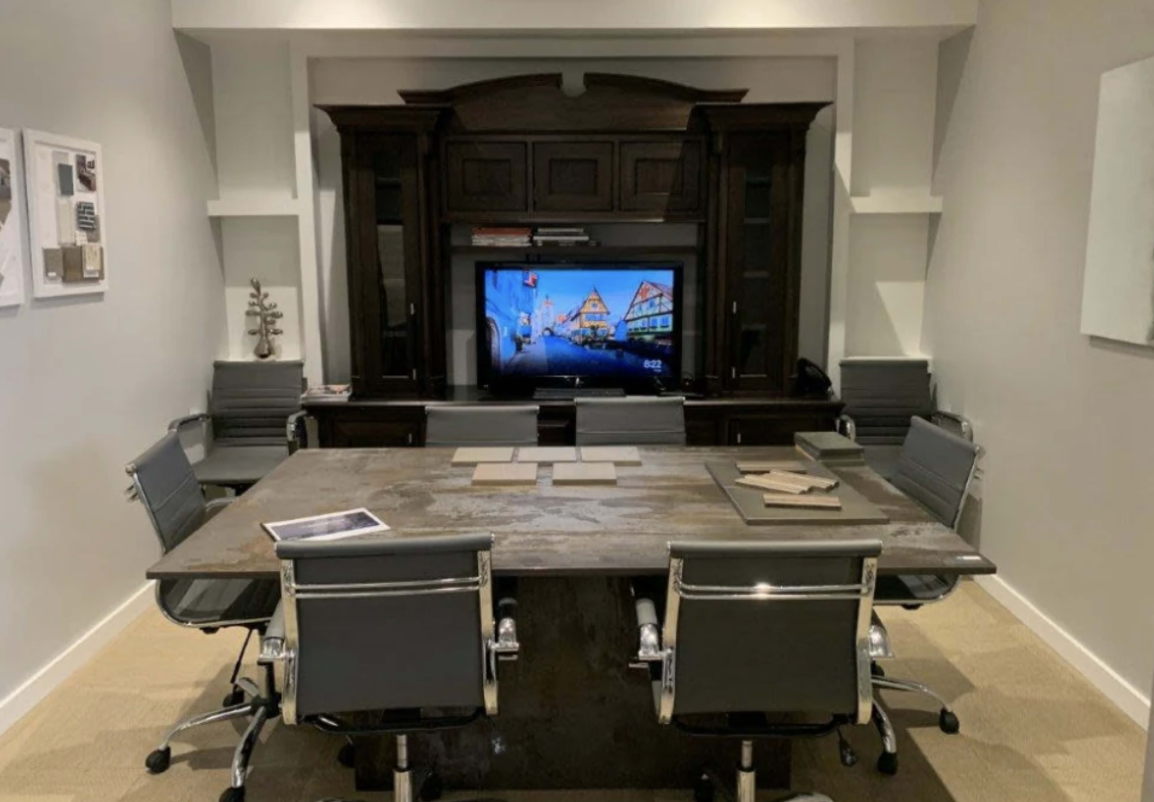 The image shows a modern conference room featuring a large wooden table surrounded by several gray office chairs and a wall-mounted television in the background.
