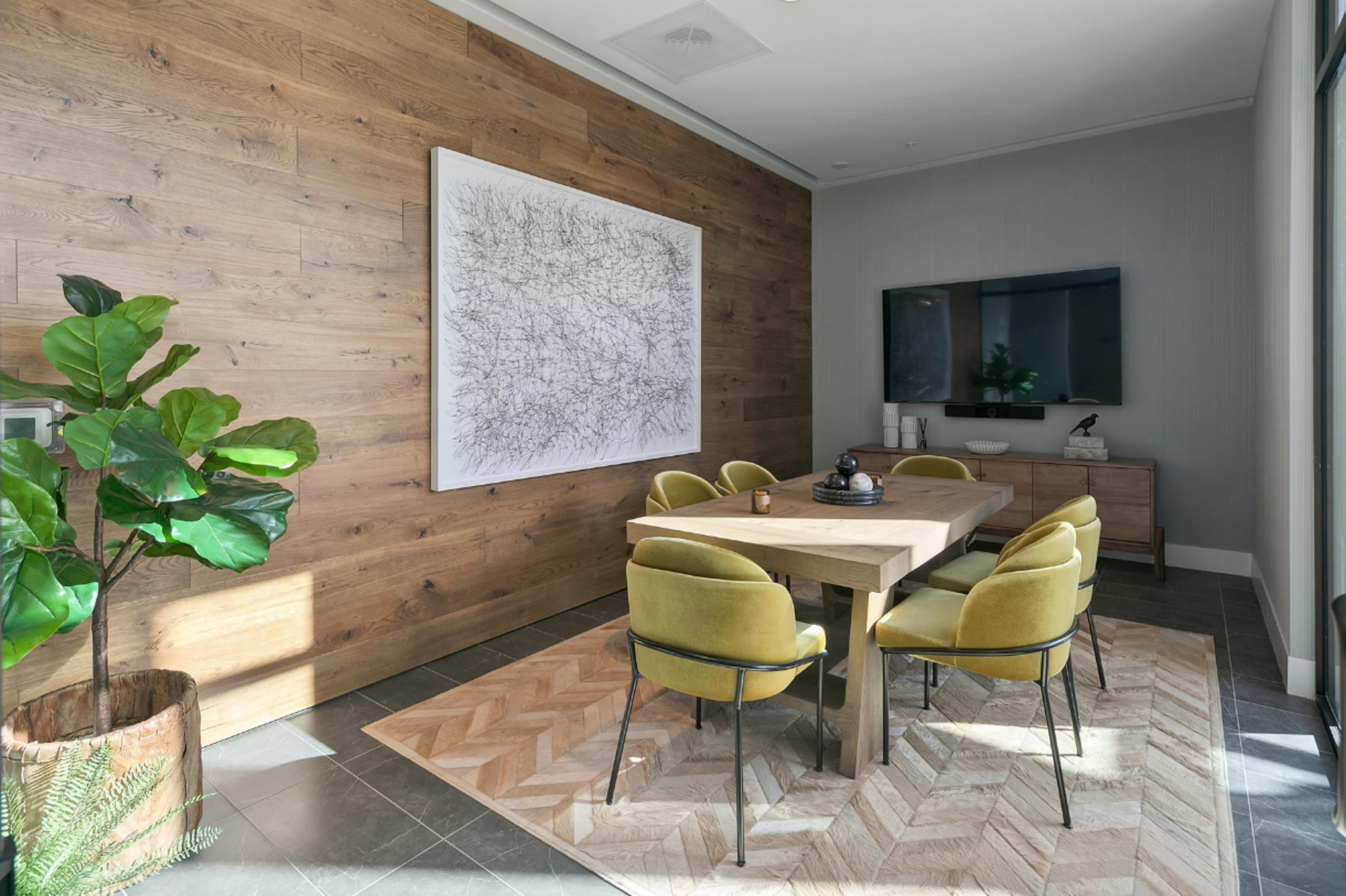 A modern dining room features a wooden table surrounded by yellow chairs, a large abstract artwork on the wall, and a television mounted above a side cabinet.