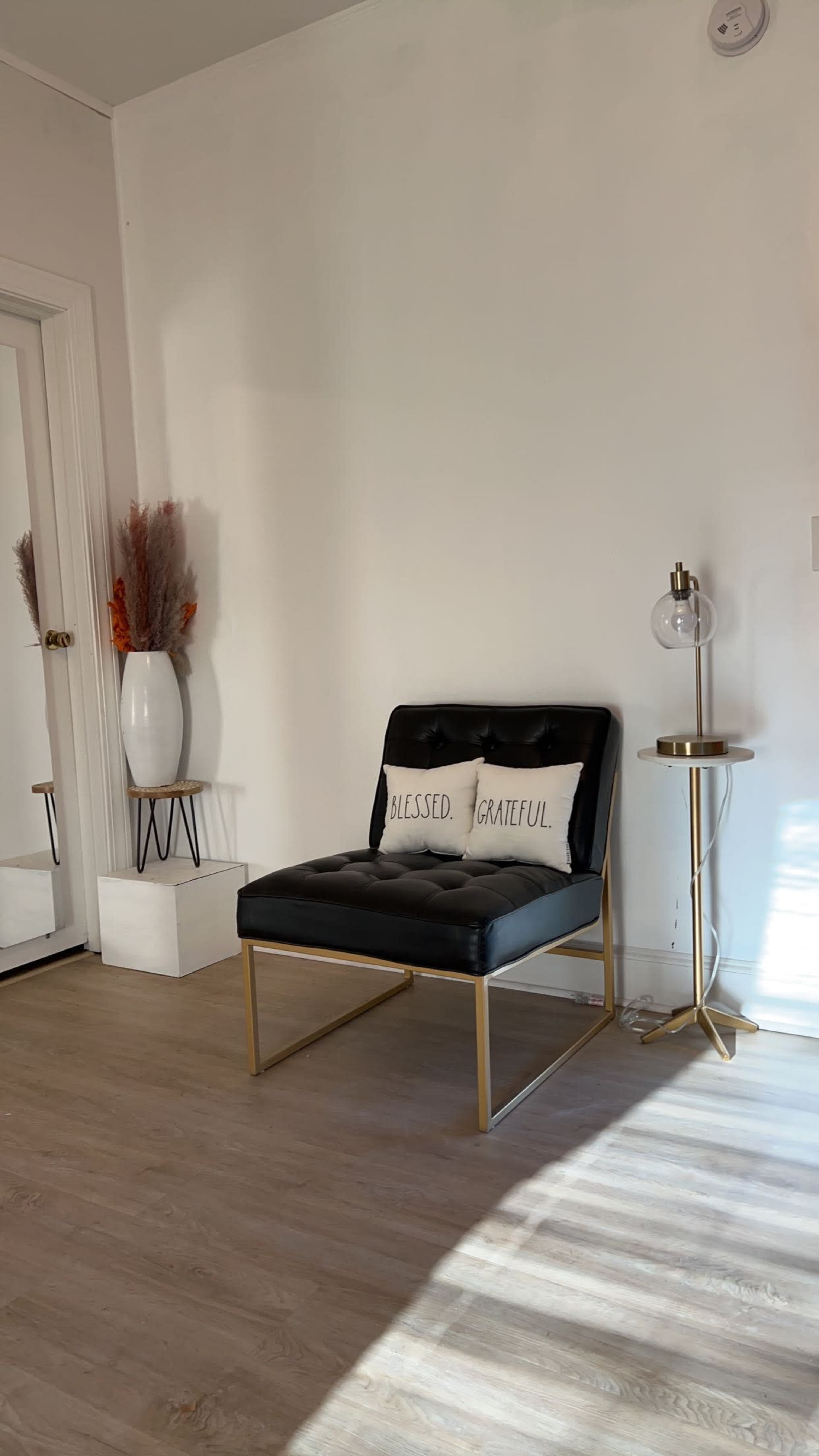 A black upholstered chair with two decorative pillows sits beside a small side table and a tall vase, against a light-colored wall.