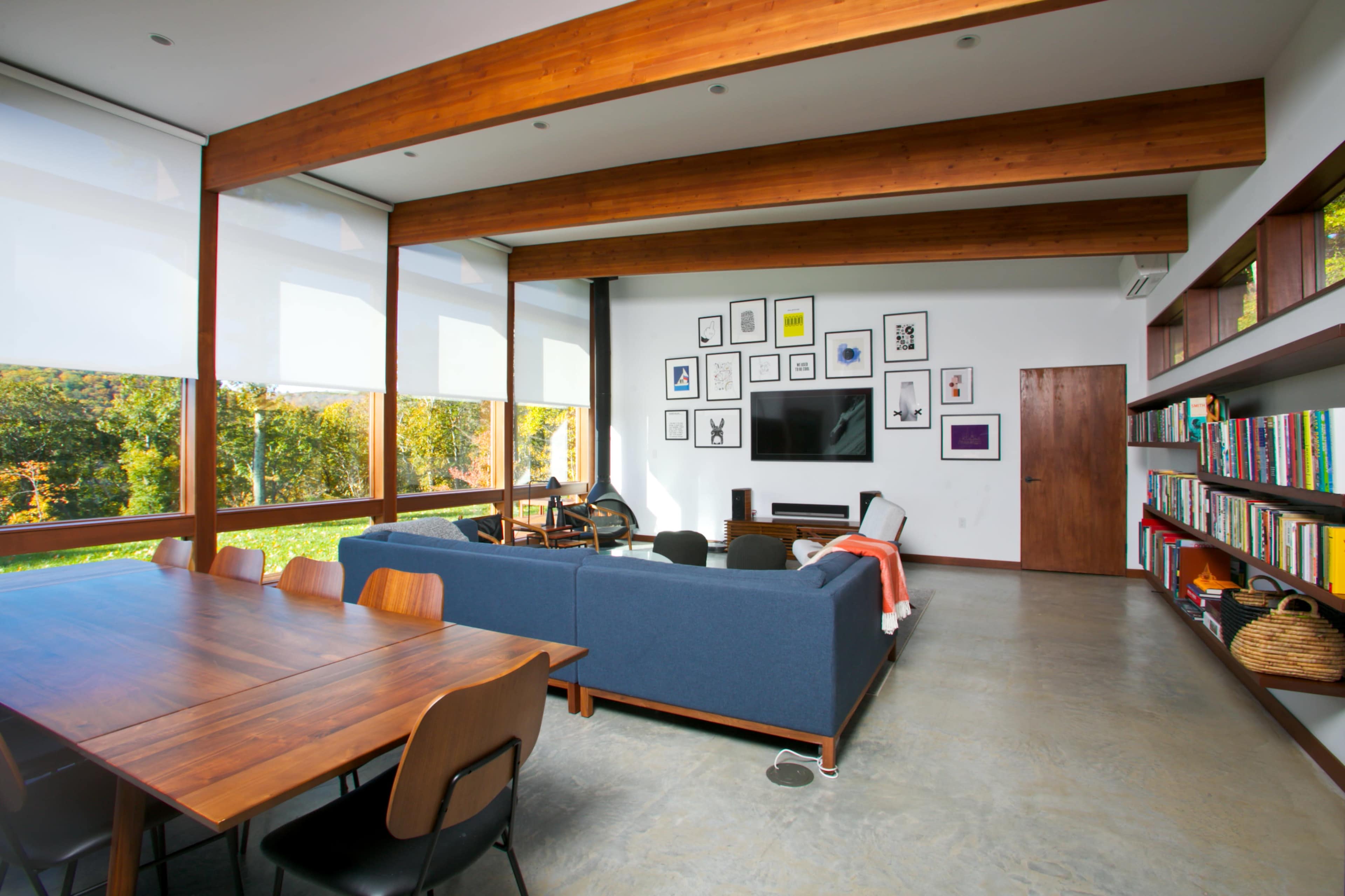 A modern living room with a blue sofa, a wooden dining table, and large windows overlooking a green landscape.