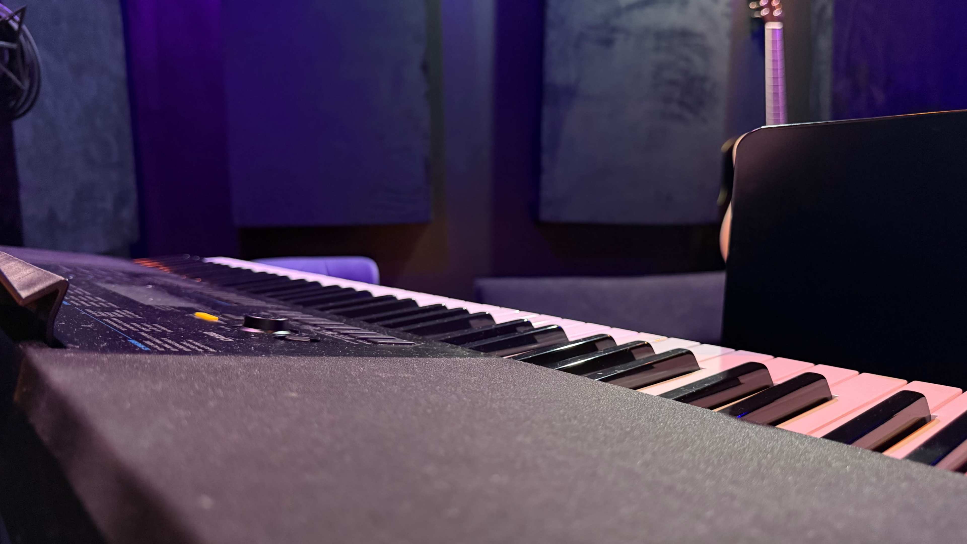 A close-up view of a keyboard with black and white keys, set against a backdrop of dark soundproofing panels and a guitar in the background.
