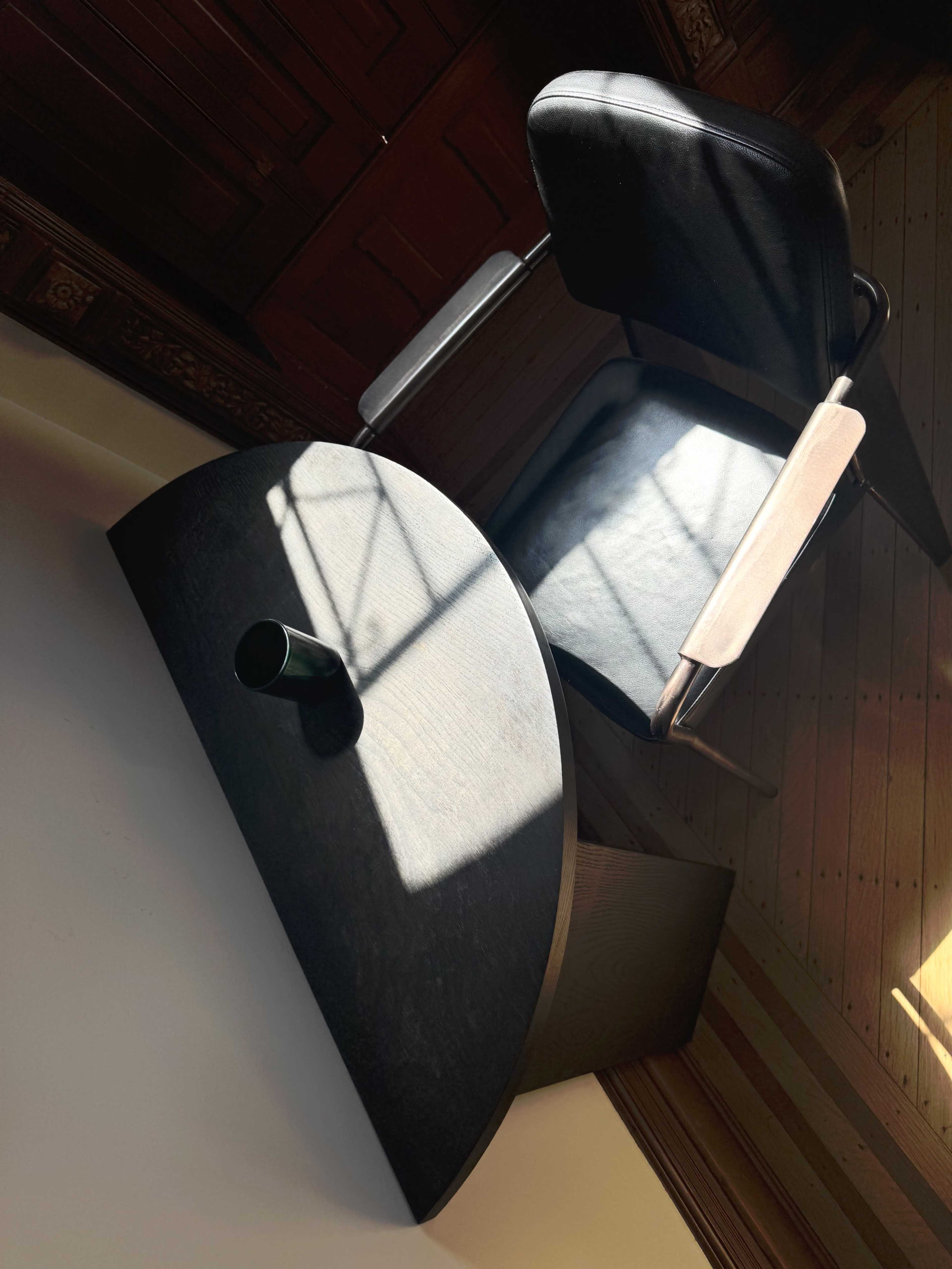 A wooden desk with a curved edge and a black chair sits against a wall, illuminated by sunlight casting shadows across the scene.