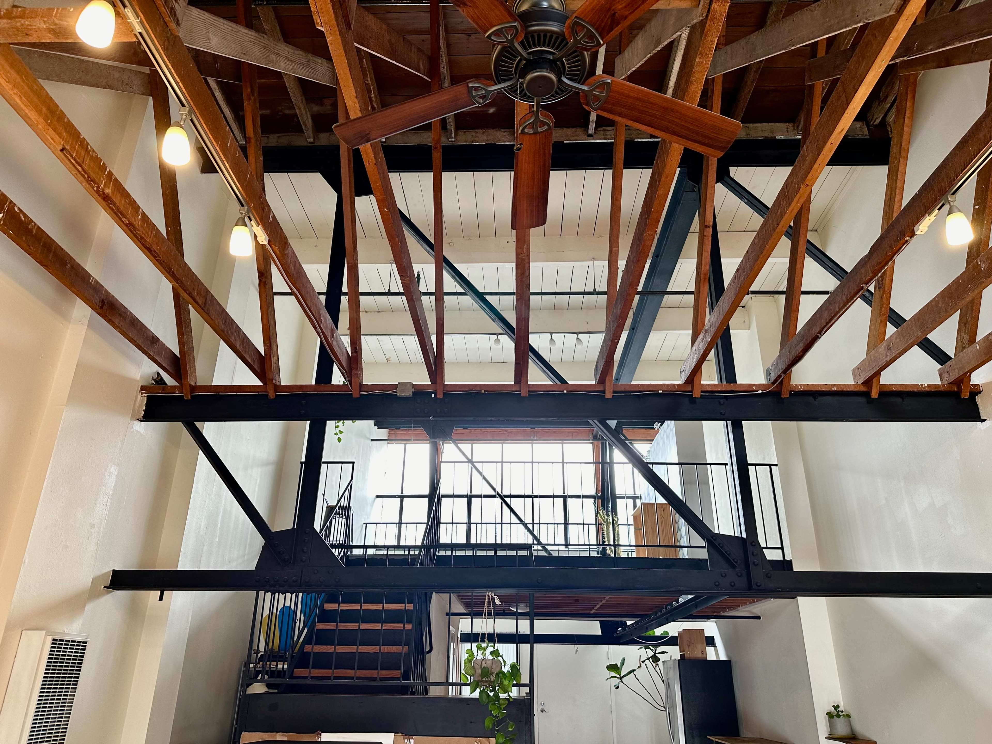 The image shows a spacious interior with exposed wooden beams and a large ceiling fan, featuring a staircase and a second-level balcony overlooking the main area.