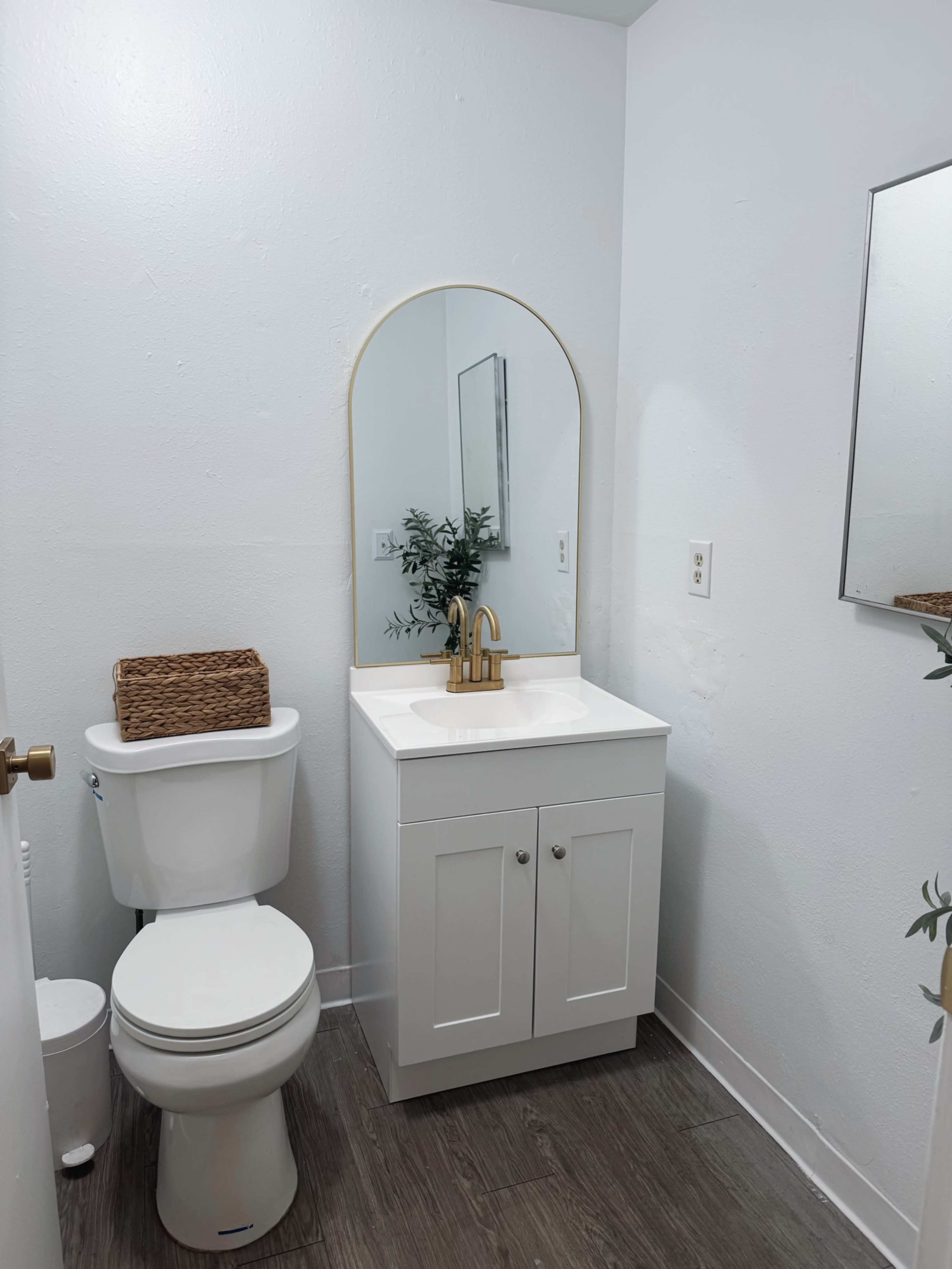 The image shows a small bathroom with a white vanity, a round mirror, a toilet, and a basket on the countertop, all set against a light-colored wall.