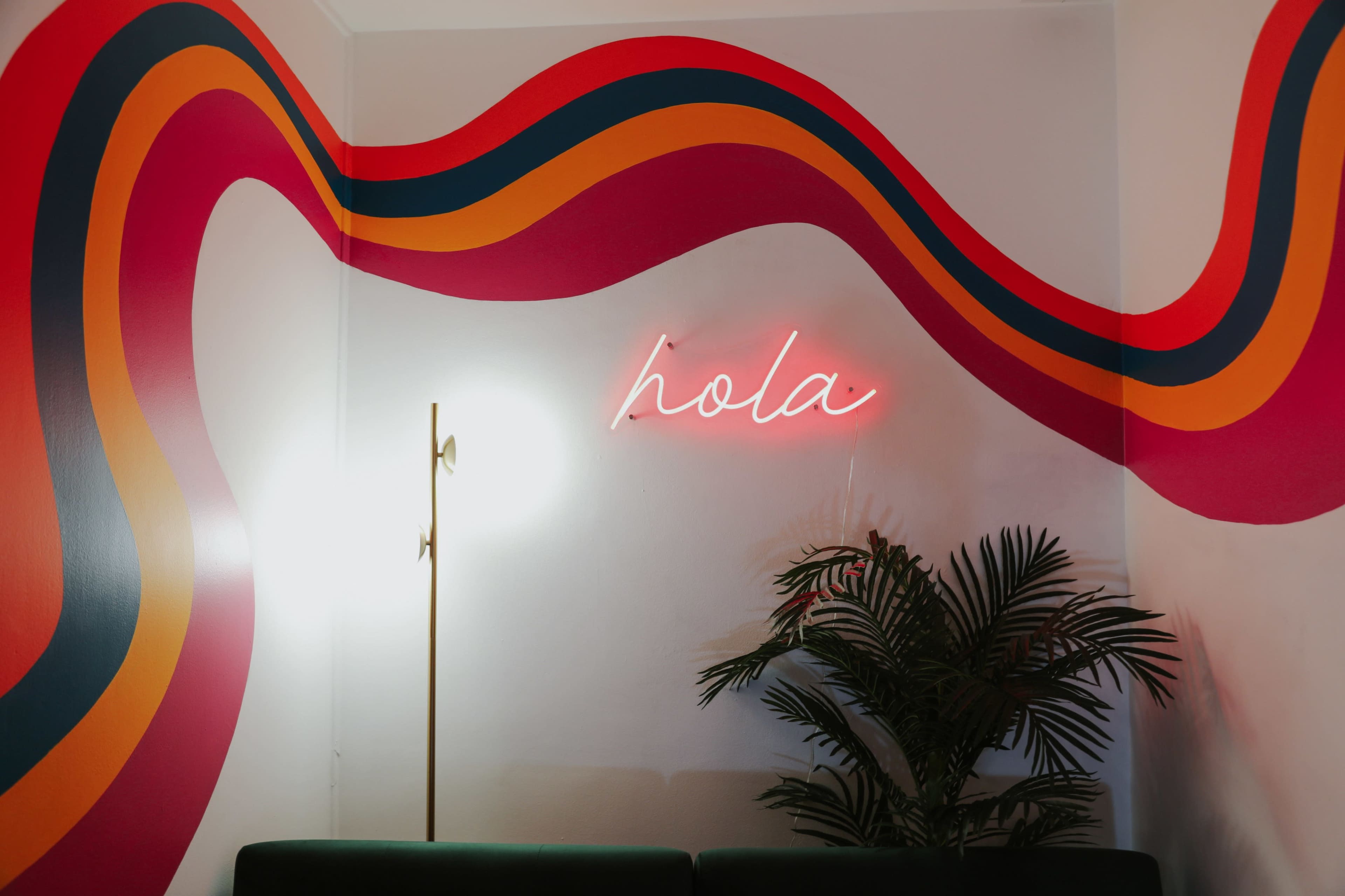 A brightly painted wall features bold, wavy stripes in red, orange, and yellow, with a neon sign that reads "hola," and a floor lamp beside a potted plant.
