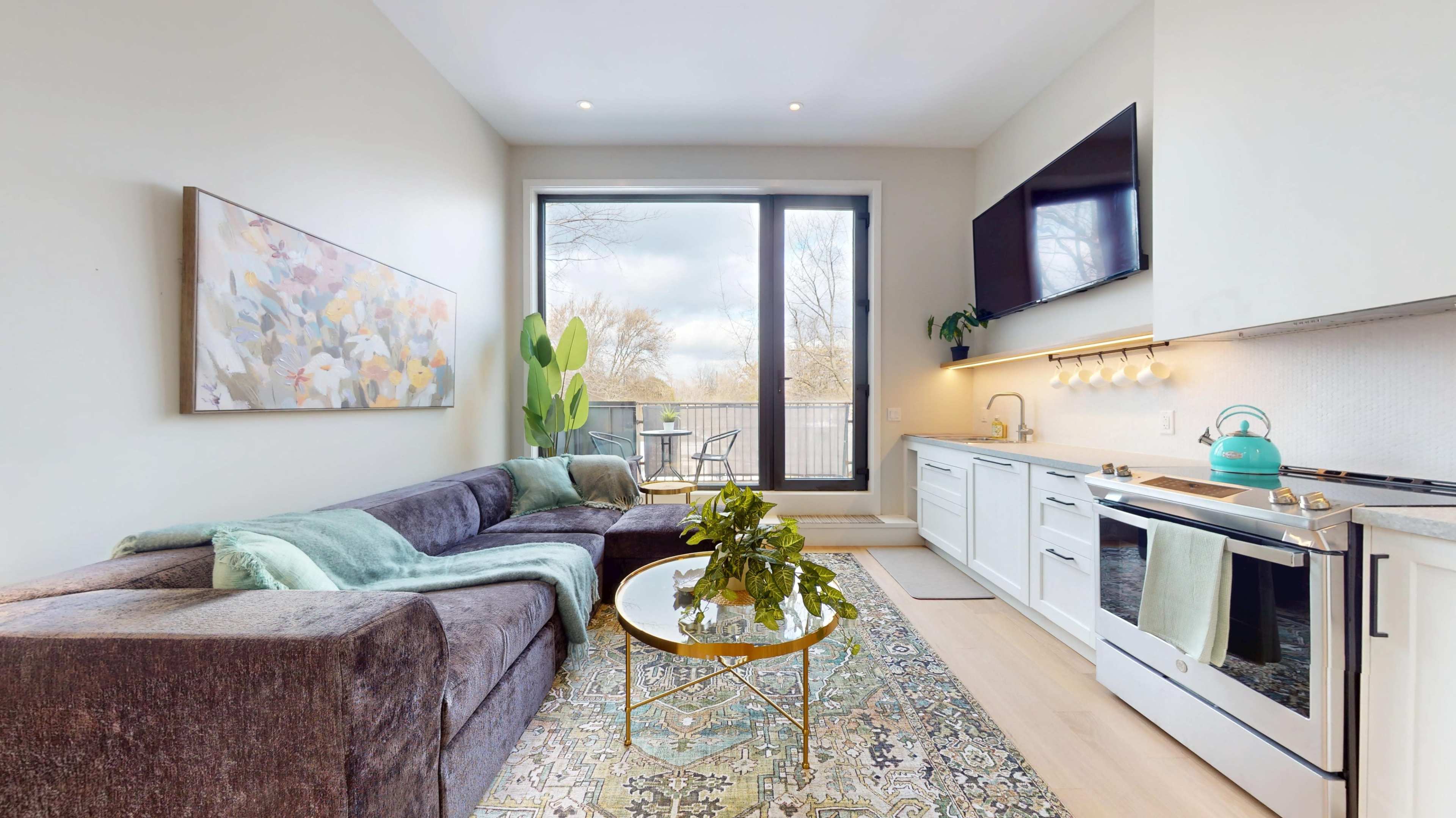 The image shows a modern living room with a gray sectional sofa, a round glass coffee table, and a kitchen area, all illuminated by natural light from large windows.