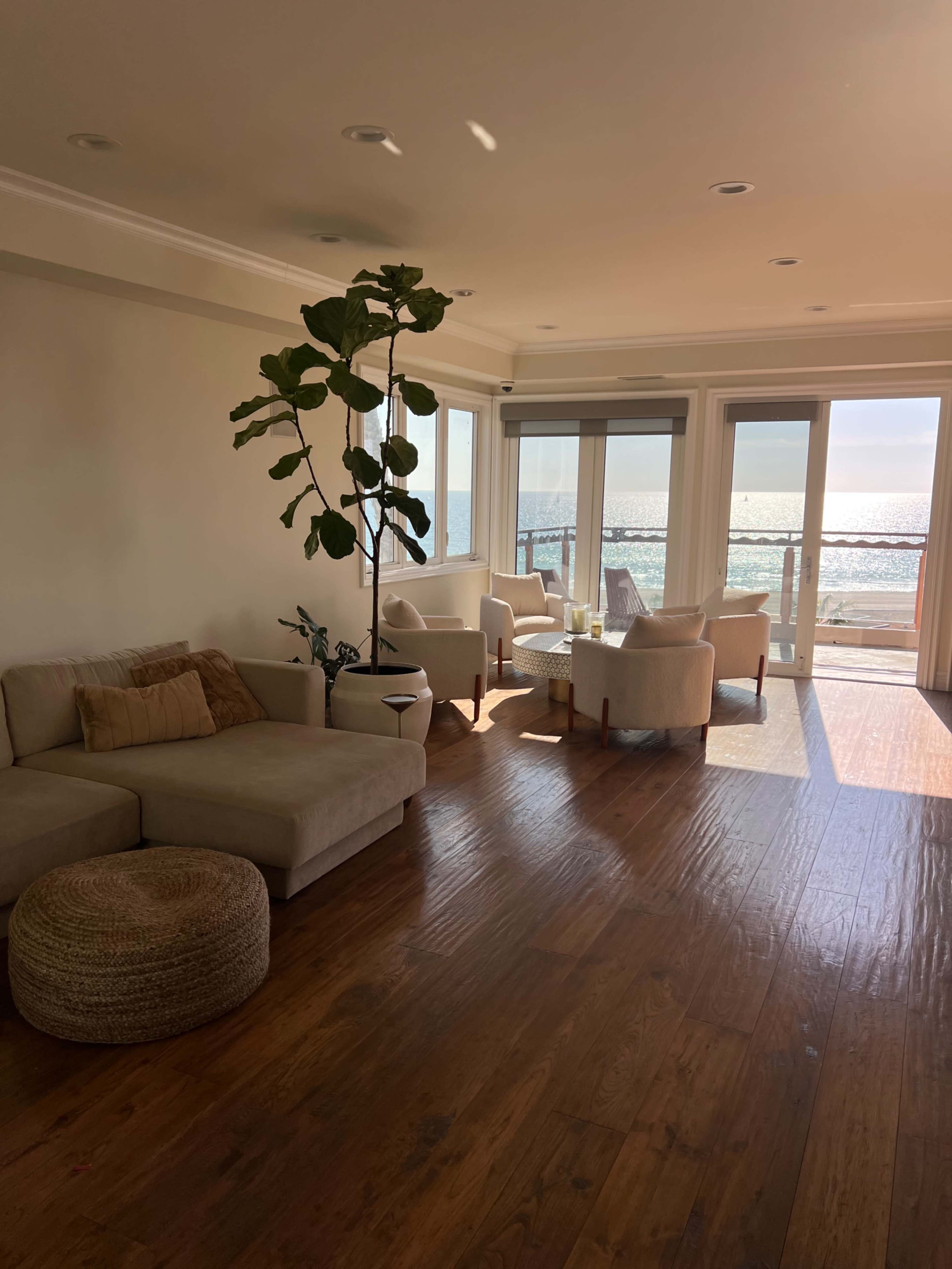 A spacious living room features a light-colored sofa, a round ottoman, and several chairs arranged near large windows overlooking the ocean.