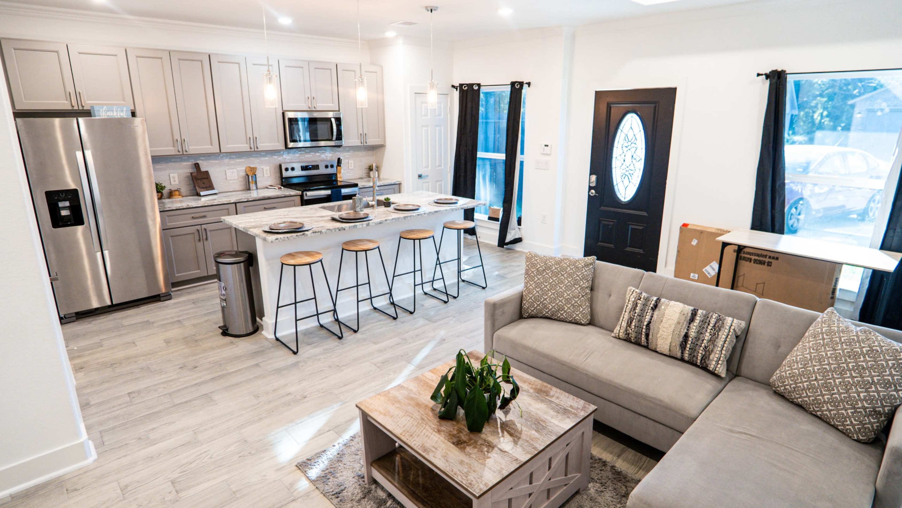 The image shows a modern kitchen and living room area featuring a grey sofa, a wooden coffee table, and a kitchen island with bar stools.