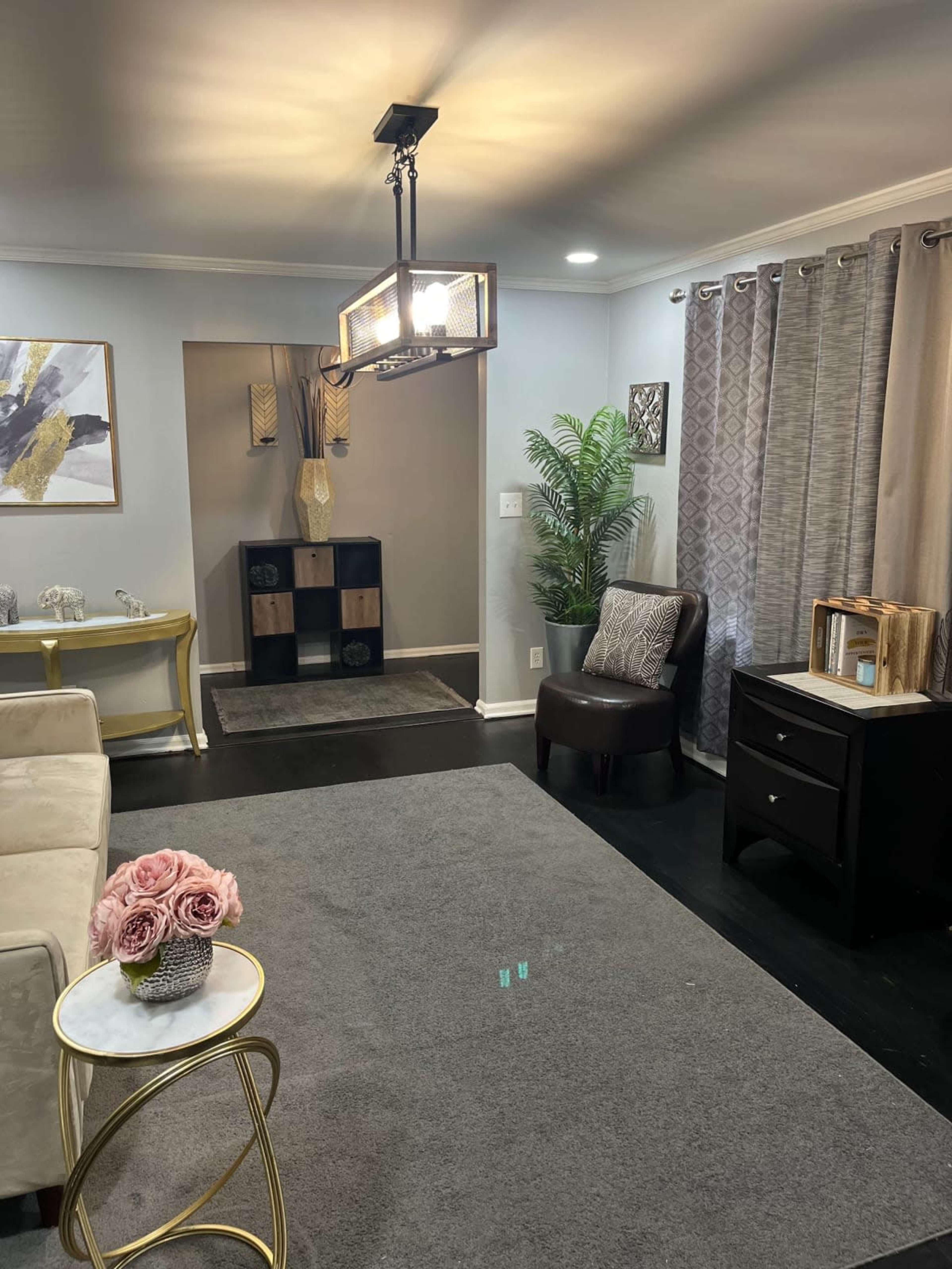 The room features a modern seating area with a light fixture, a small table with flowers, and decorative elements including curtains and a plant.