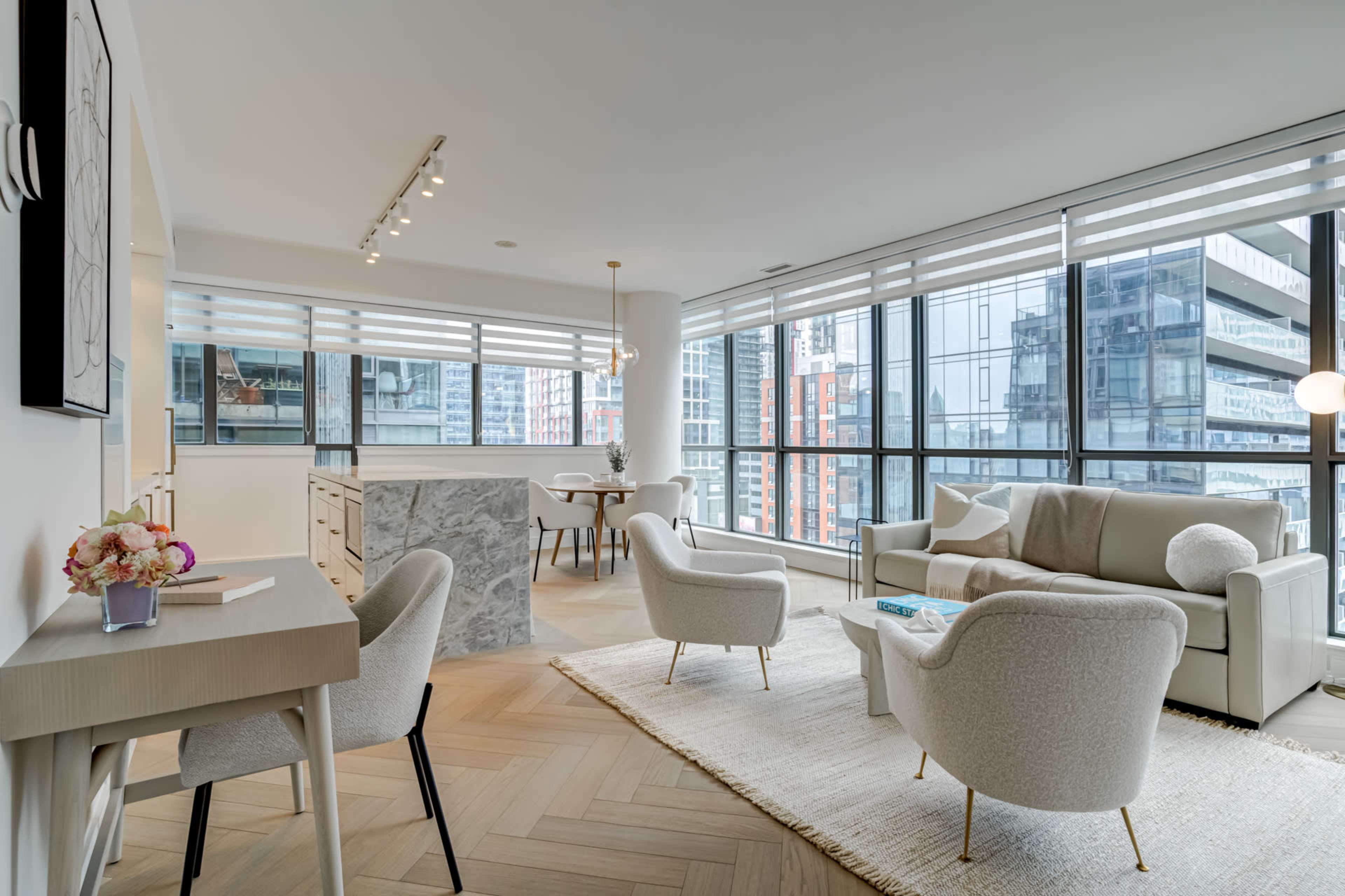 A modern living room features a light-colored sofa, two armchairs, and large windows overlooking city buildings.