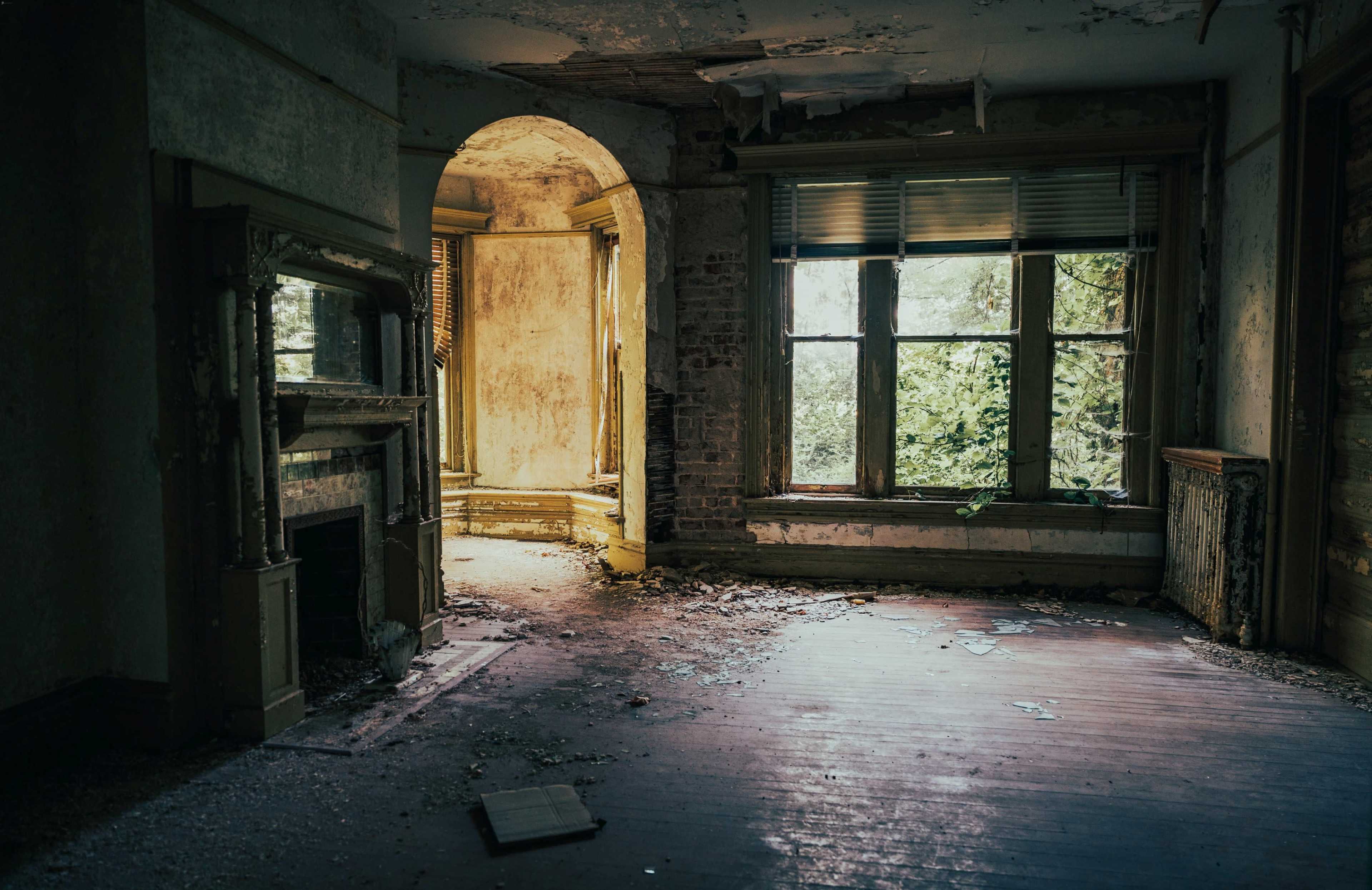 An abandoned room with peeling walls, broken furniture, and sunlight streaming through a window overgrown with vegetation.